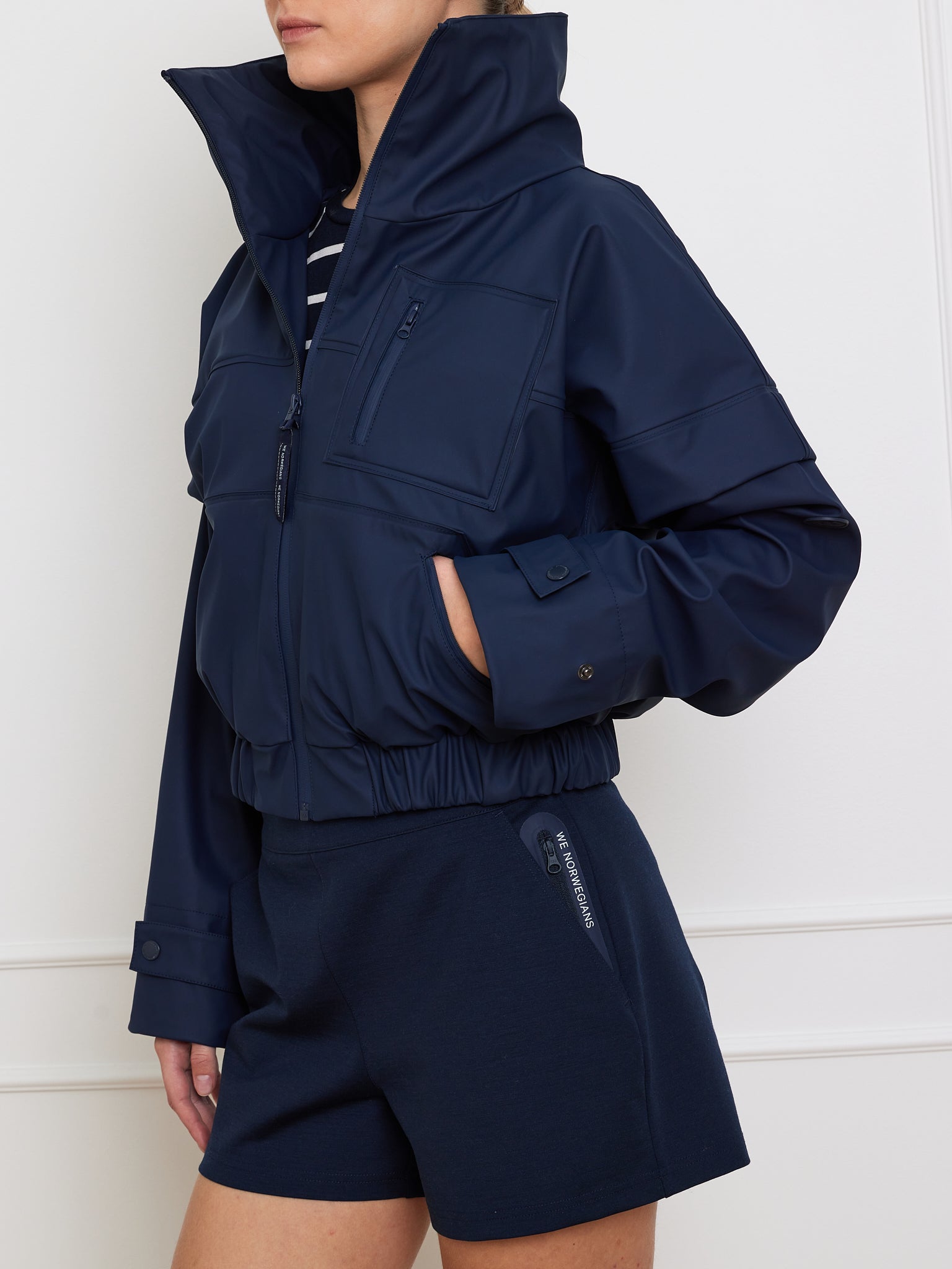 Wearing We Norwegians' Bergen Rain Jacket Women with 80s flair, a high collar, and waterproof zippers, they pair it with navy shorts and stand confidently against a plain white wall.