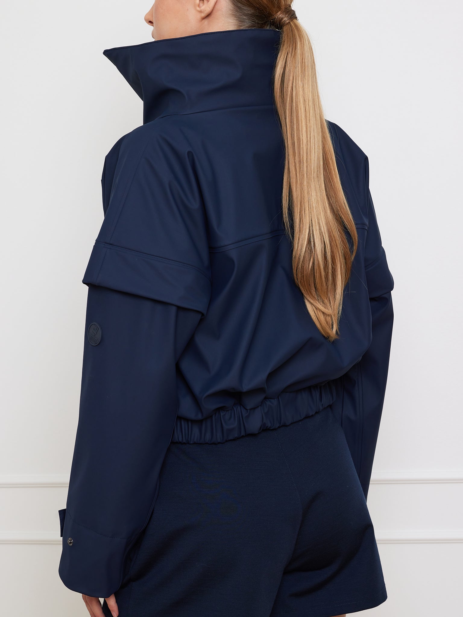 A person with long blonde hair is wearing the Bergen Rain Jacket Women by We Norwegians. It displays an 80s style with waterproof zippers and an elastic waistband, in a dark blue hue featuring a high collar and matching shorts, set against a neutral background.