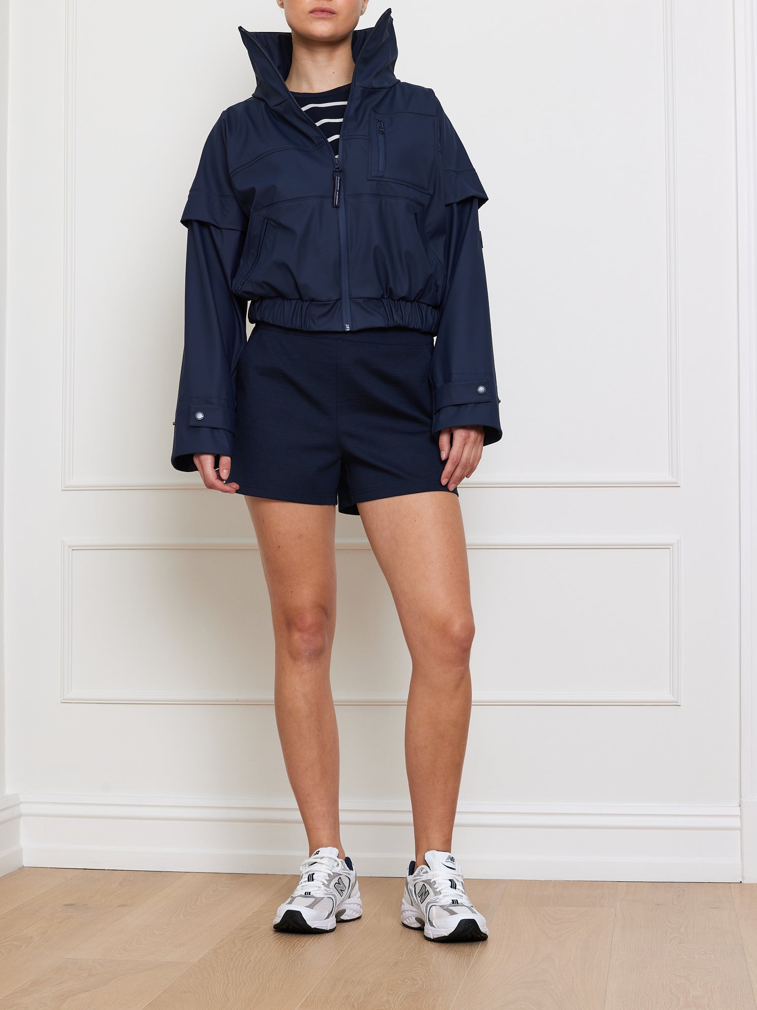 Wearing a Bergen Rain Jacket Women by We Norwegians, navy shorts, a striped top, and white sneakers on a light wooden floor with a white paneled backdrop, this person channels an effortless 80s style.