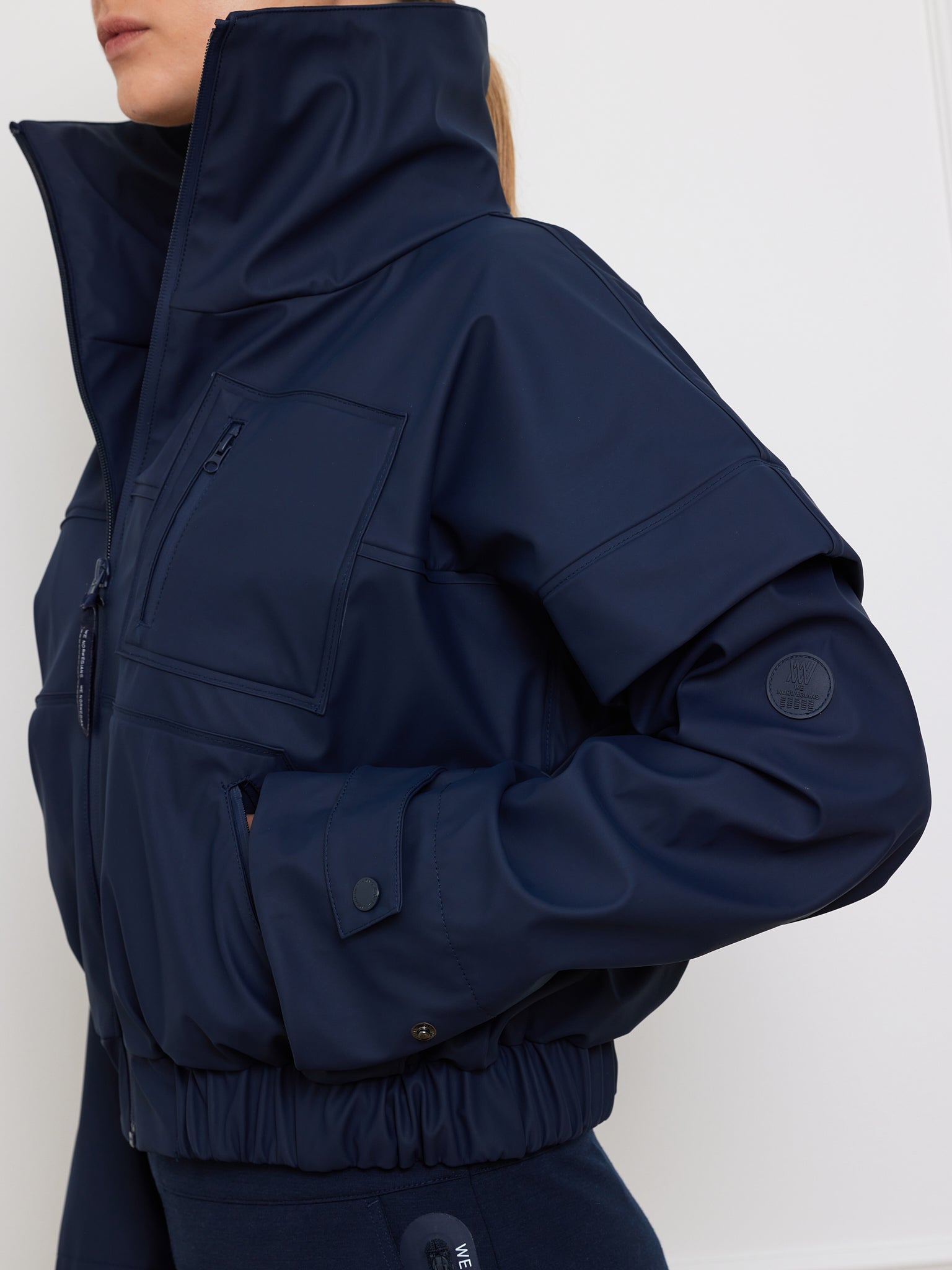 A person in the We Norwegians Bergen Rain Jacket Women, featuring a dark blue hue, high collar, pocket with logo patch on the sleeve, elastic waistband, snap button wrists, and waterproof zippers against a plain white wall.