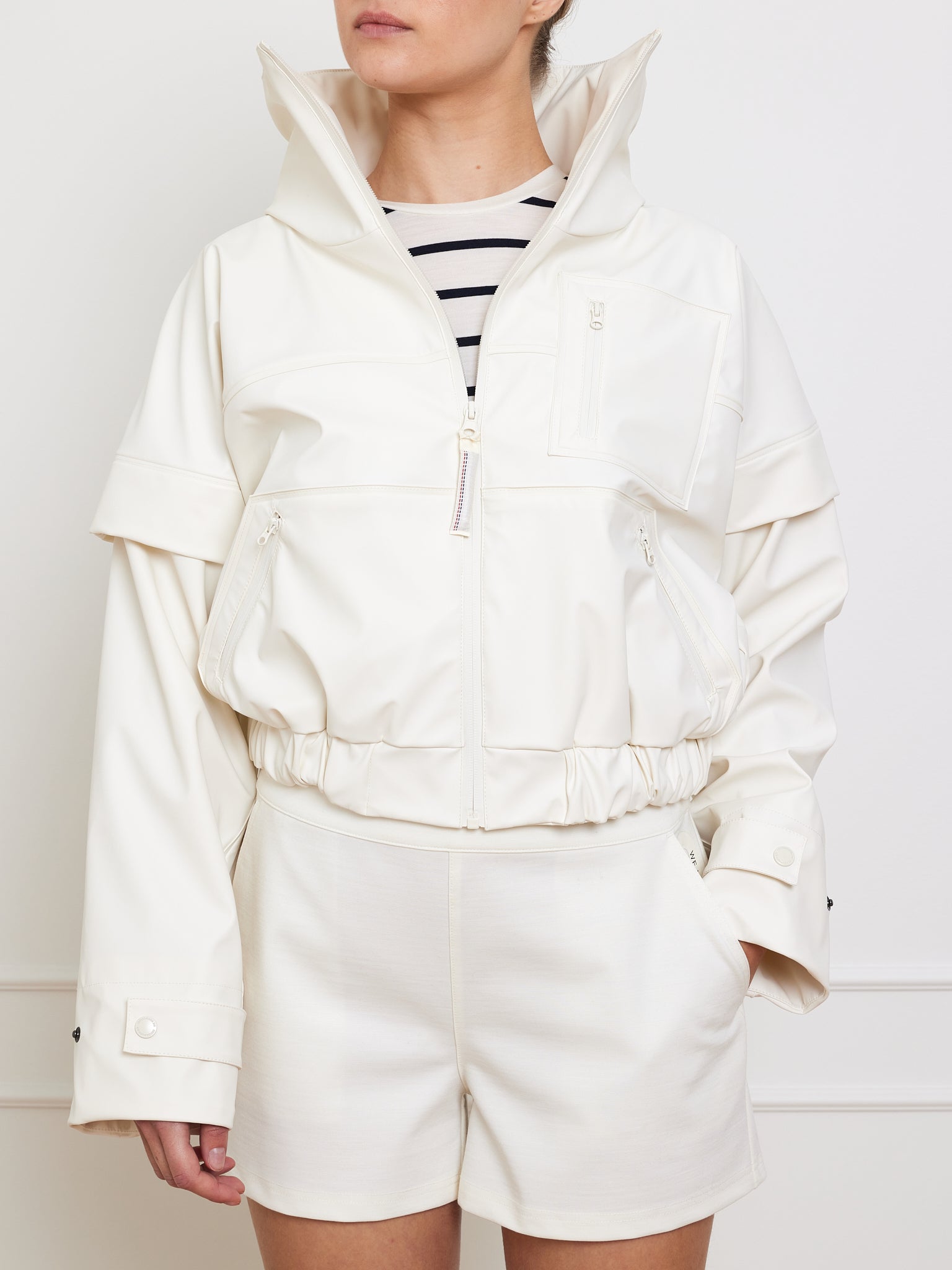 Someone channels an 80s vibe with the We Norwegians Bergen Rain Jacket Women, featuring waterproof zippers, layered over a black-and-white striped top and complemented by white shorts. Their left hand casually rests in the shorts pocket against a plain white backdrop.