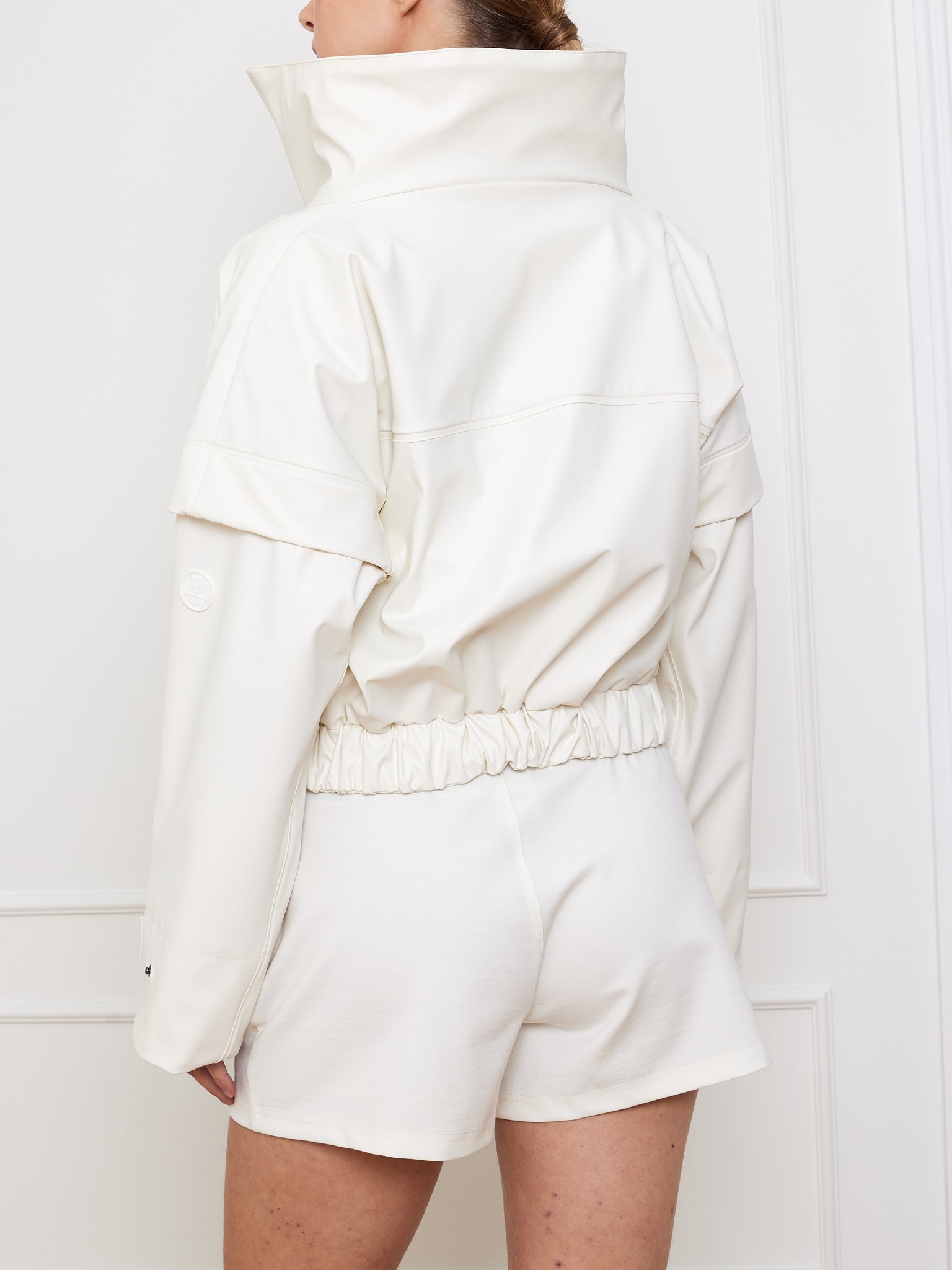 A person in a high-collared white Bergen Rain Jacket Women by We Norwegians, paired with loose white shorts, stands facing away. The jacket has an 80s vibe with waterproof zippers and an elastic waistband against a plain light wall.