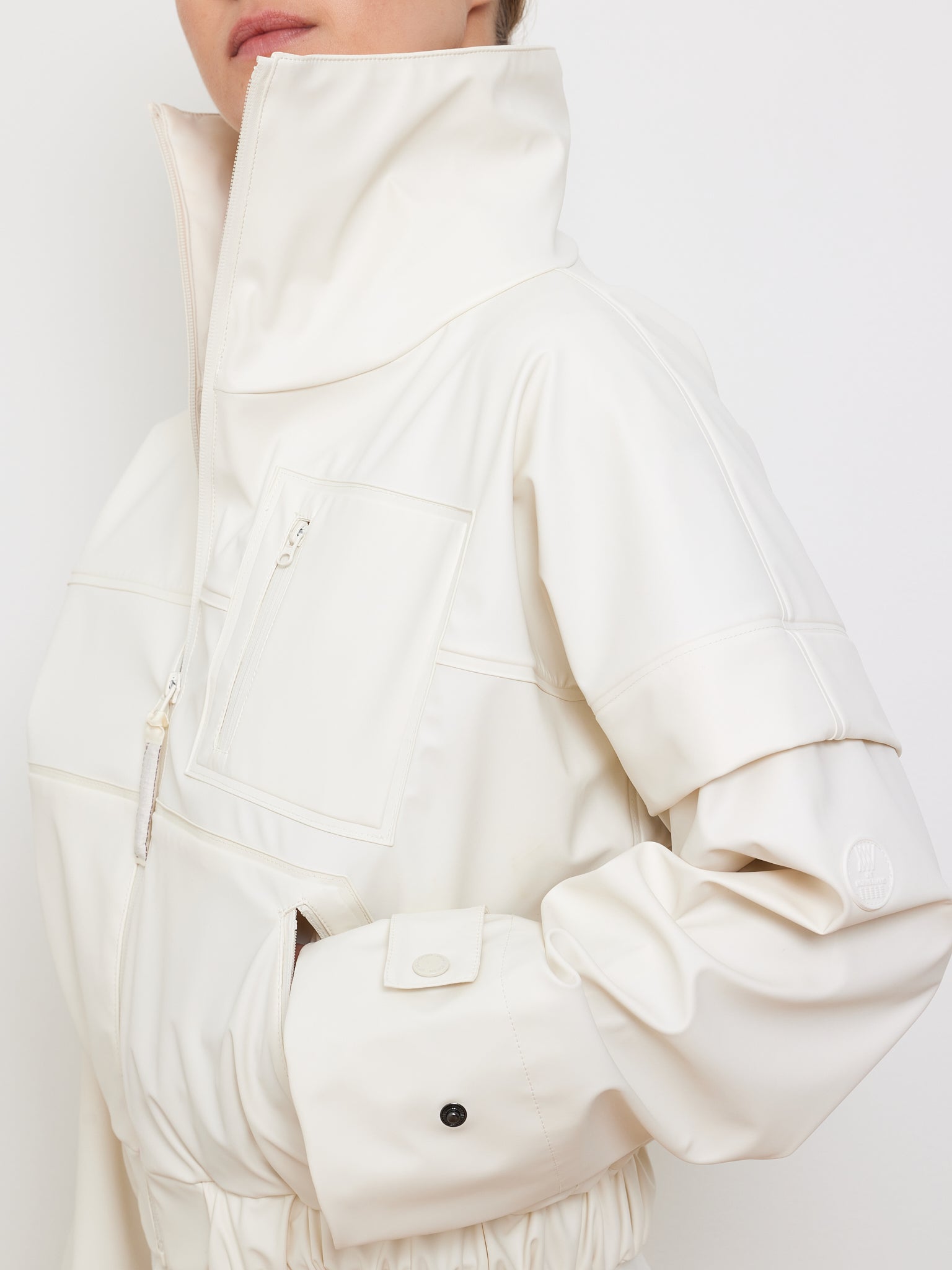 A model showcases We Norwegians' Bergen Rain Jacket Women, featuring an 80s style high-collar, white design with waterproof zippers, an elastic waist, and buttoned cuffs. The hand rests casually in a side pocket against a plain white backdrop.