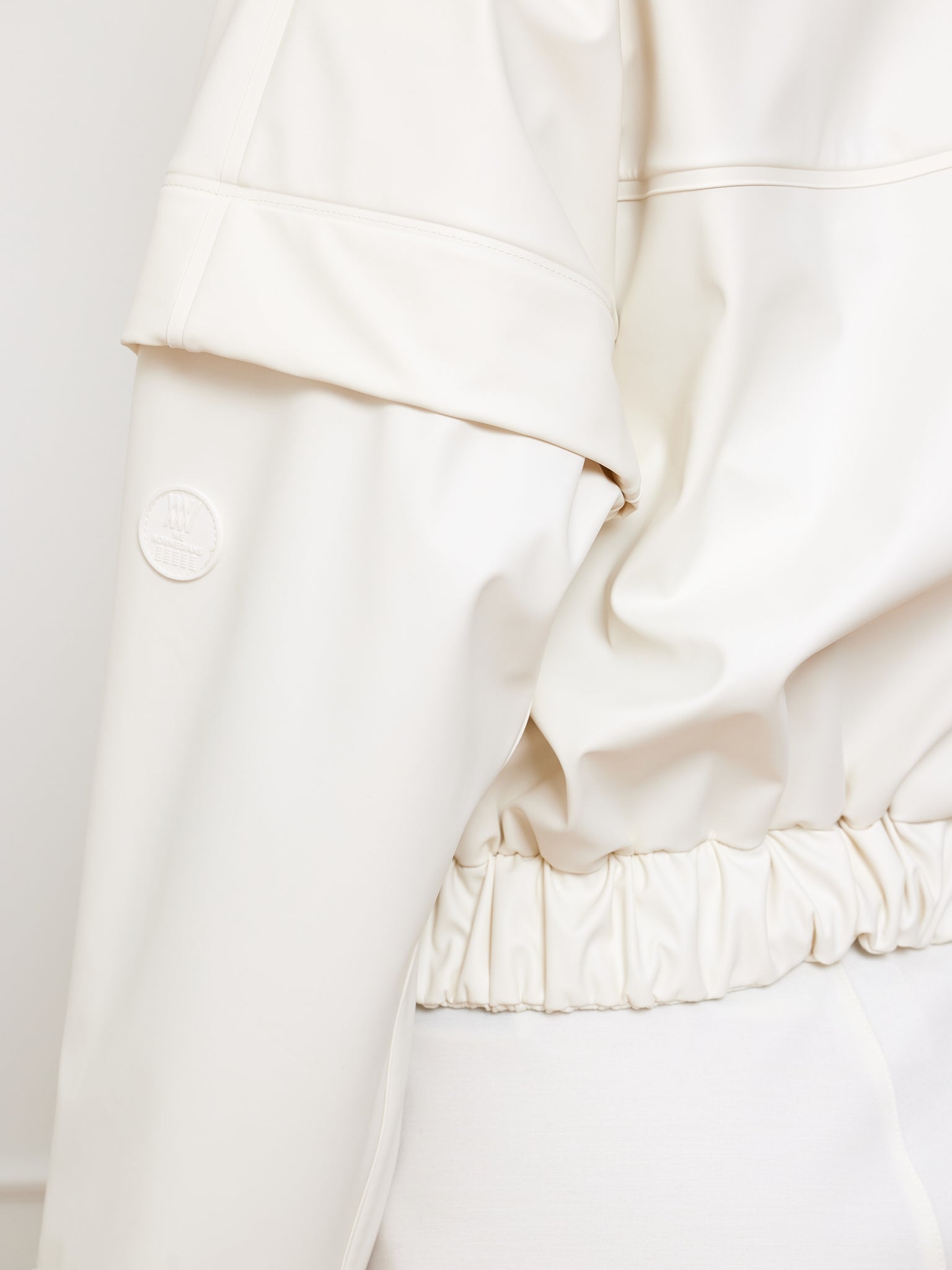 A close-up of the Bergen Rain Jacket Women by We Norwegians shows a cream-colored bomber with an 80s glossy finish. The back and sleeve reveal gathered elastic at the hem, waterproof zippers, and a round logo on the arm, all set against a plain white background.
