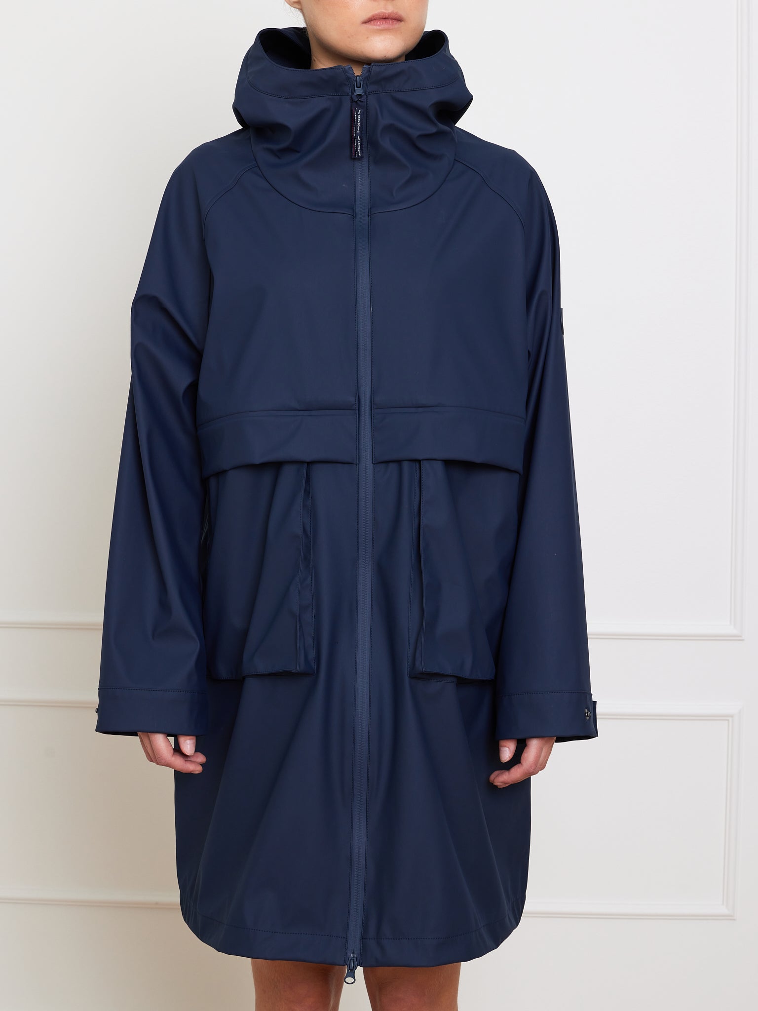 Someone dons a We Norwegians Bergen Rain Coat Women in dark blue, oversized with a hood and zipper front. Large pockets and a layered design ensure waterproofing. The face is hidden as they stand against a plain white wall.