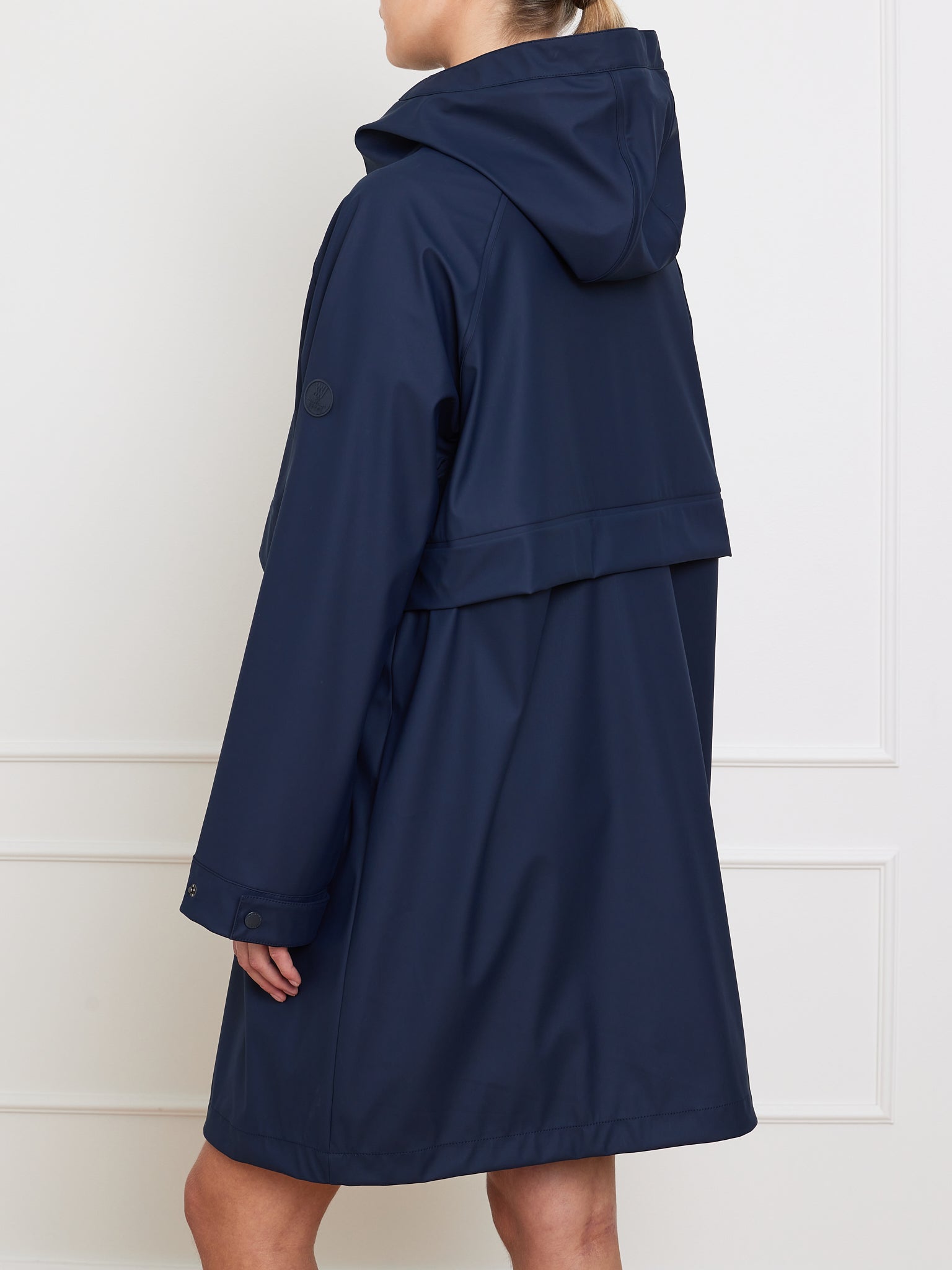 A person models the We Norwegians Bergen Rain Coat Women in navy blue against a white background. The oversized, waterproof coat has long sleeves and back flap detail. The individual is shown from the side, facing away from the camera.