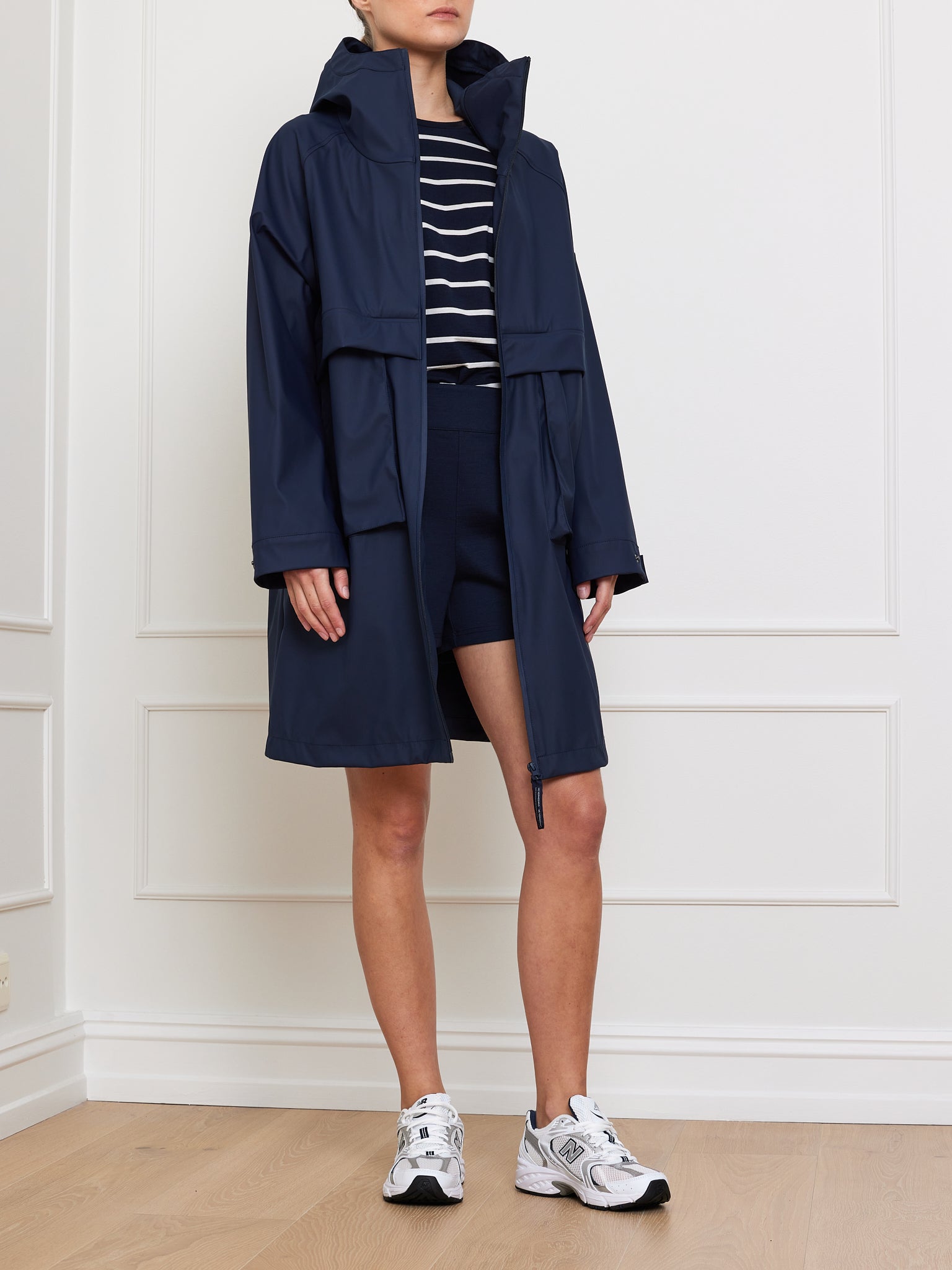 Modeling the We Norwegians Bergen Rain Coat Women in navy, styled with a striped shirt, black shorts, and white sneakers. Set against a white paneled wall on a wooden floor, this oversized fit raincoat perfectly showcases its sleek waterproof design as the individual stands with hands by their sides.