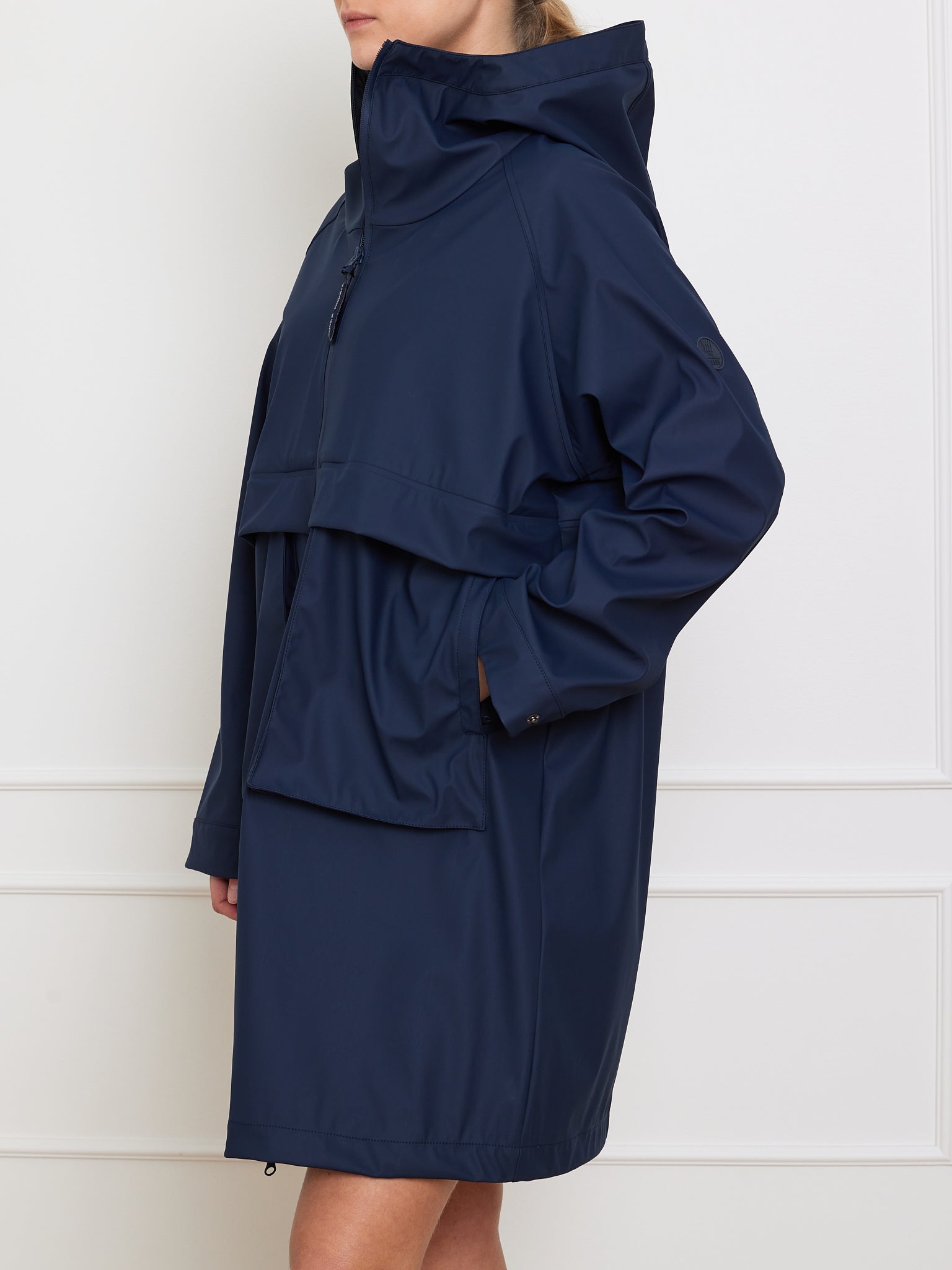 Someone is wearing We Norwegians' navy blue Bergen Rain Coat Women, which has a high collar, oversized fit, and waterproof front pockets. They're standing side profile against a plain white wall with their hands tucked in the pockets.