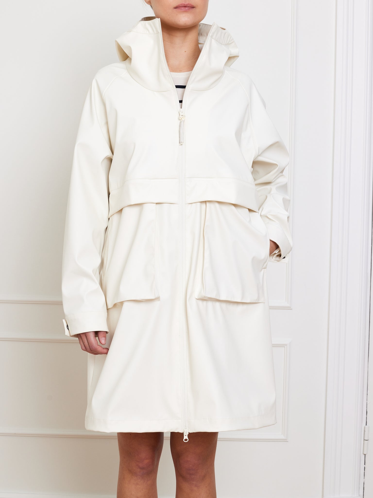 A person is seen in a Bergen Rain Coat Women by We Norwegians, oversized with a hood and long white design. It has large front pockets, layered style, and a front zipper. They stand with one hand in the pocket against a white paneled wall; their face remains unseen.