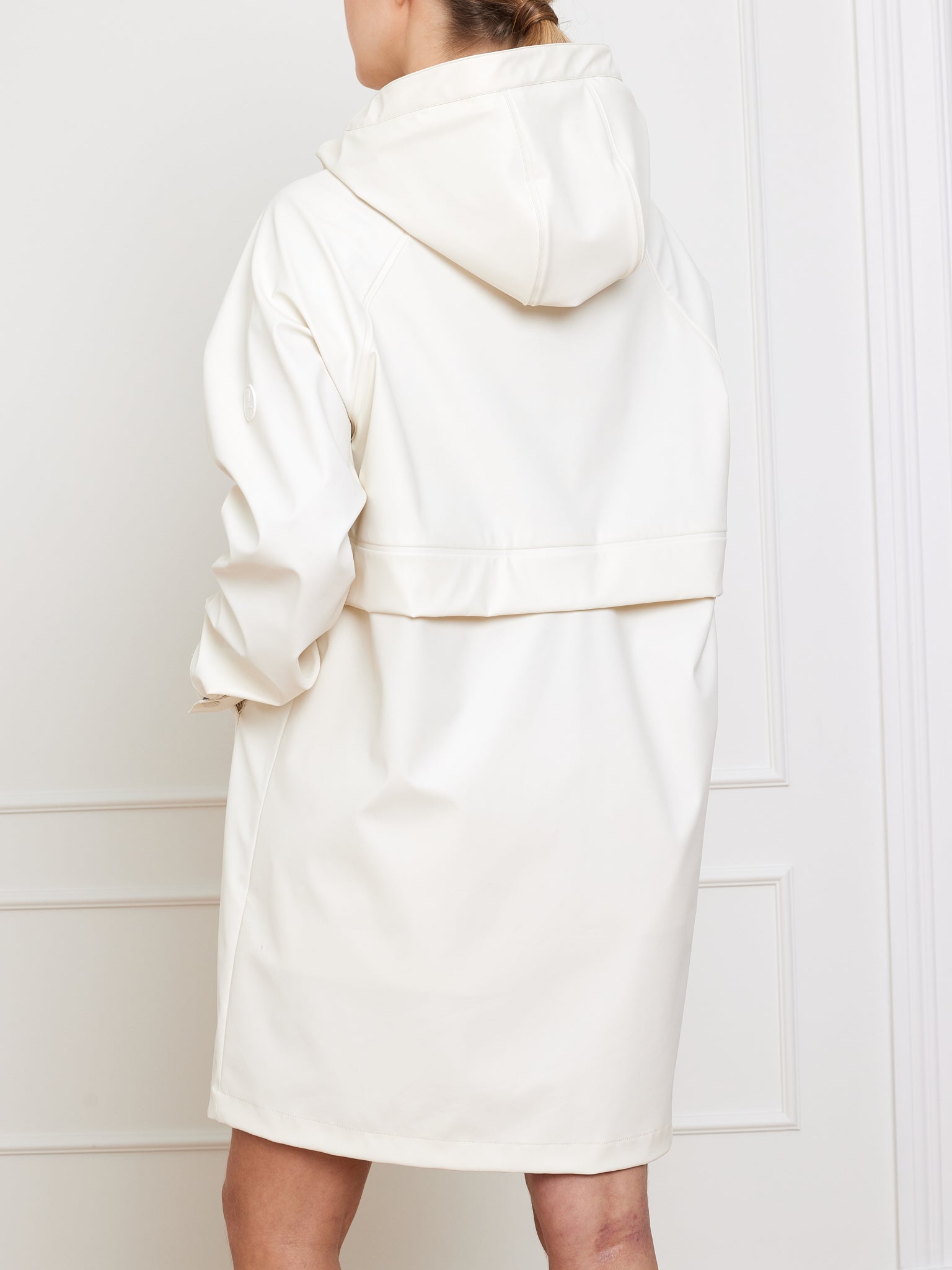 A person is standing indoors against a white paneled wall, facing away, wearing the Bergen Rain Coat Women by We Norwegians—a long, white waterproof coat with an oversized fit.