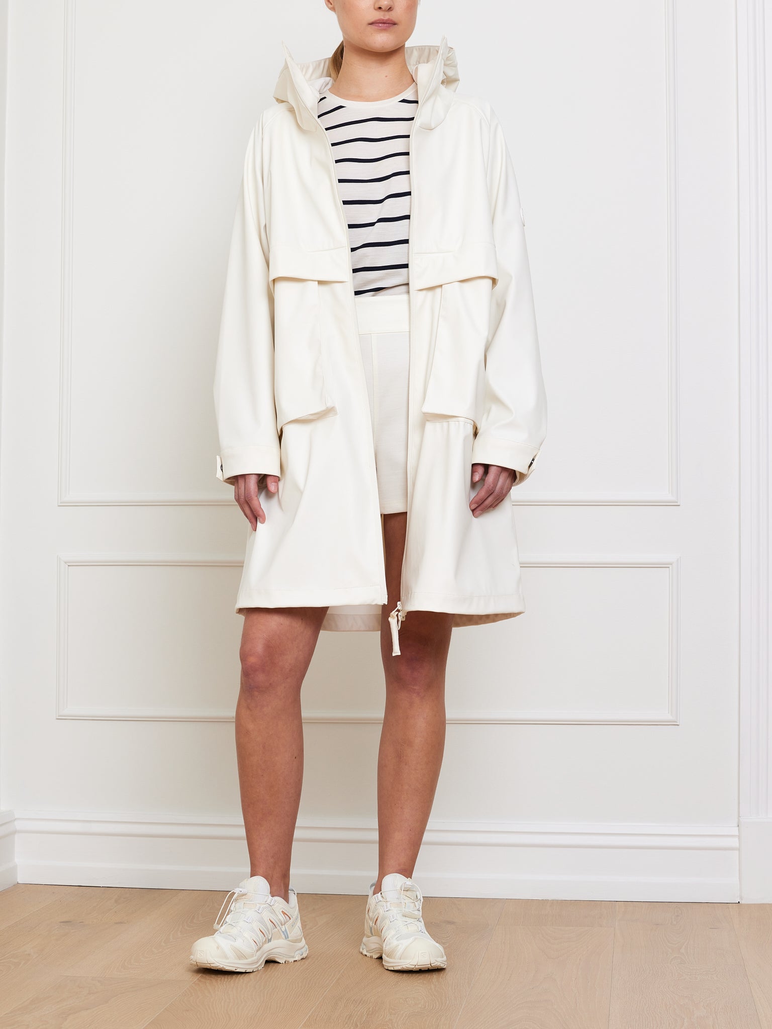 A person is in a room with white paneled walls and wooden flooring, wearing an oversized fit Bergen Rain Coat Women in cream by We Norwegians over a striped shirt and shorts, with white sneakers.