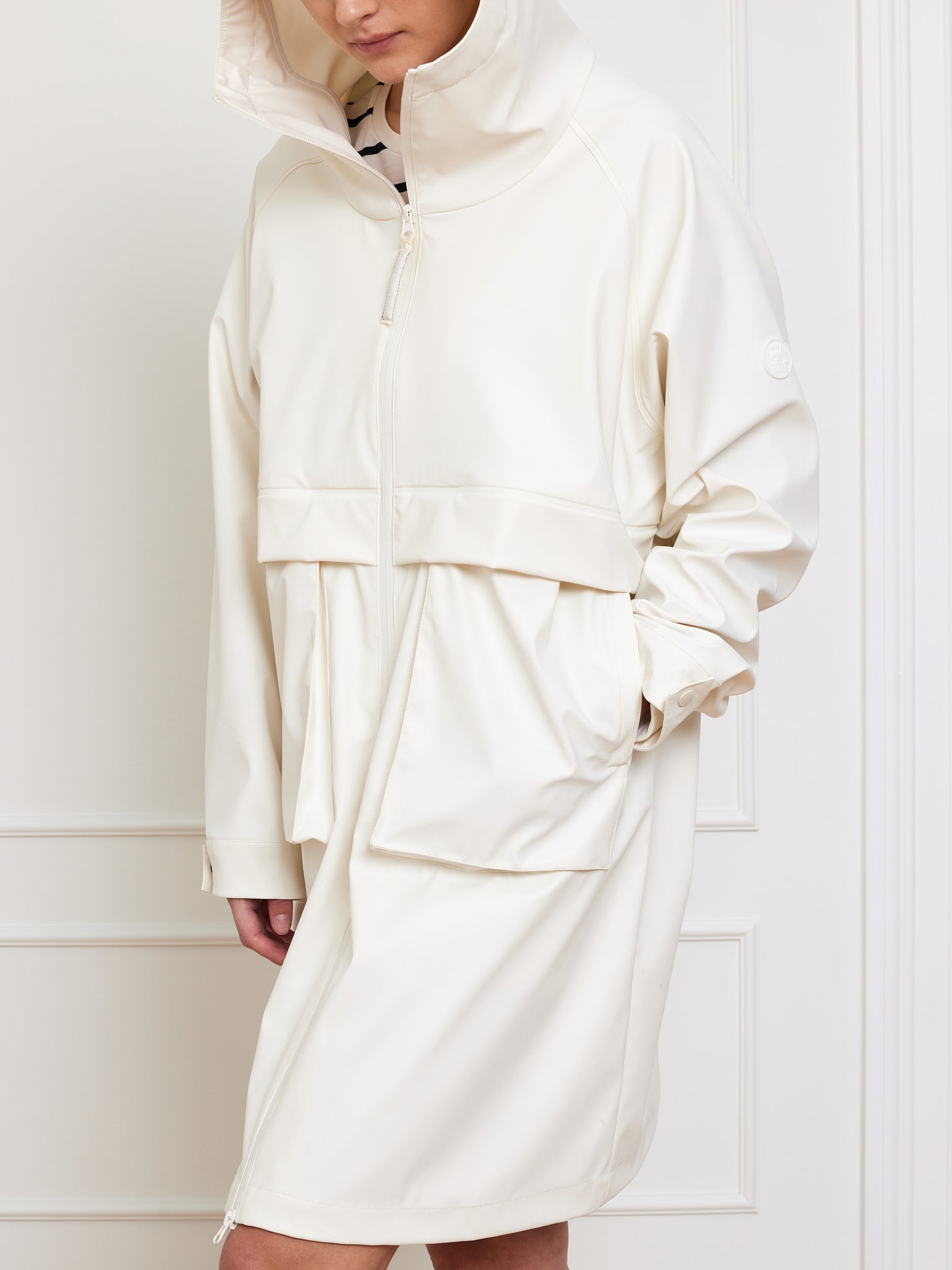 A person, face unseen, leans against a white wall wearing an off-white Bergen Rain Coat Women by We Norwegians. It's knee-length with a sleek, minimalist design, high collar, and large pockets. The oversized fit adds allure as hands rest casually in the pockets.