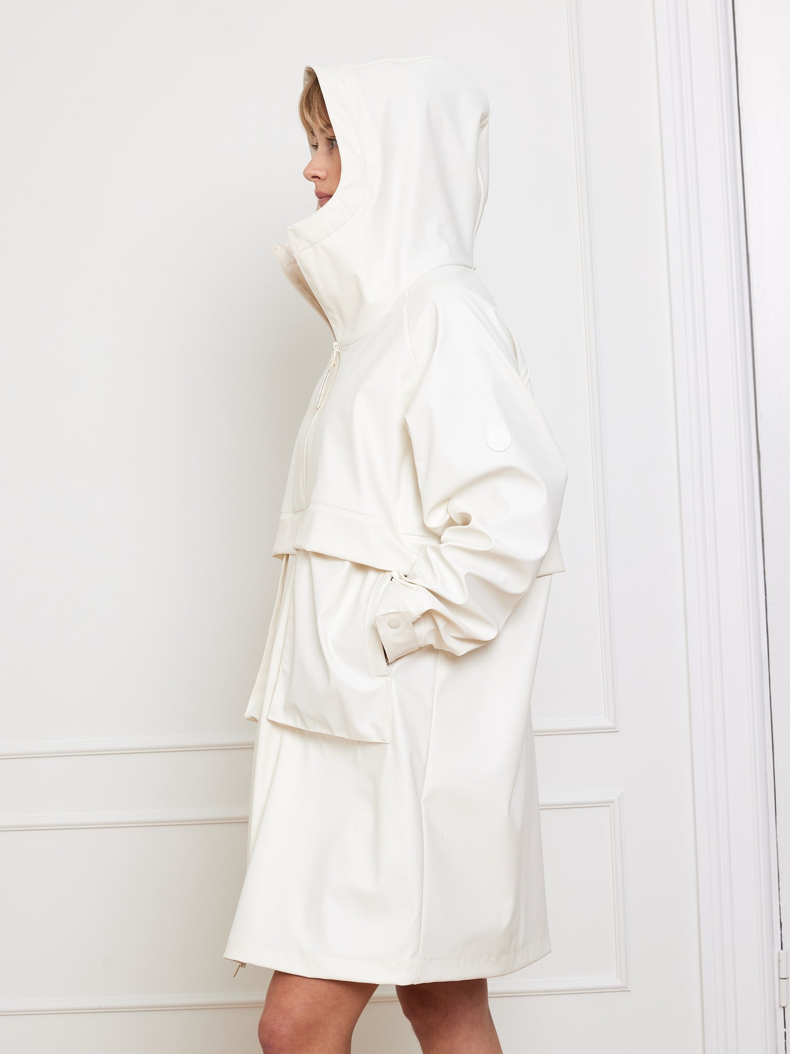 A person stands in profile against a plain wall with decorative molding, wearing We Norwegians' Bergen Rain Coat Women. The white waterproof coat showcases an oversized fit, high collar, cape-like back detail, and the wearer has their hands in pockets.
