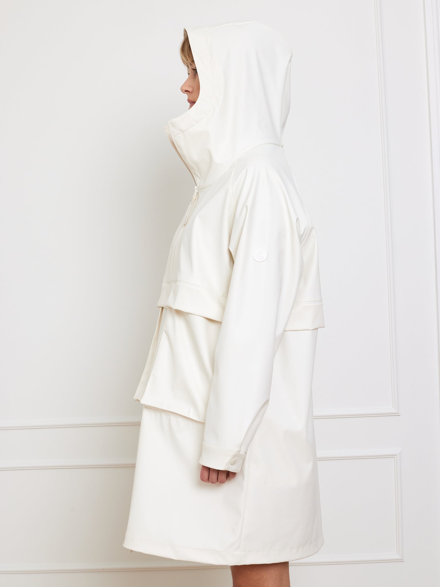 A woman wearing an oversized fit Bergen Rain Coat Women by We Norwegians, with a white hood, stands in profile against a plain white wall.