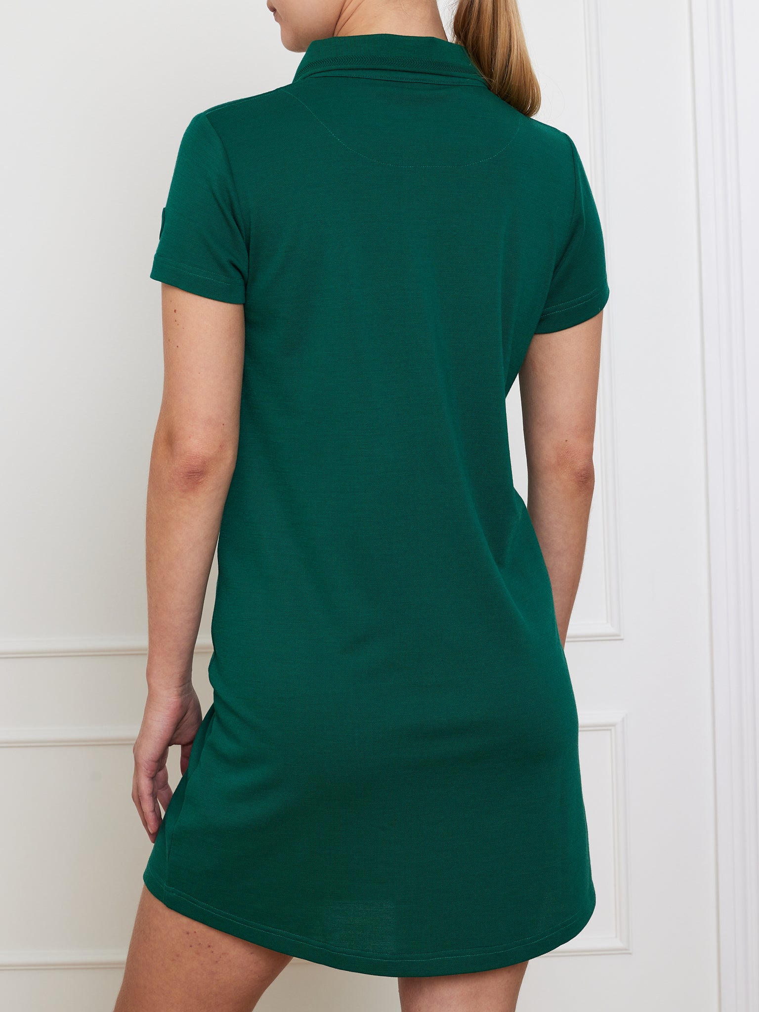Rear view of a person in a short-sleeved, dark green We Norwegians Medalist Piqué Dress Women. The collared, loose-fit dress provides moisture-wicking comfort. The person's long hair is tied back as they stand against a white paneled wall.