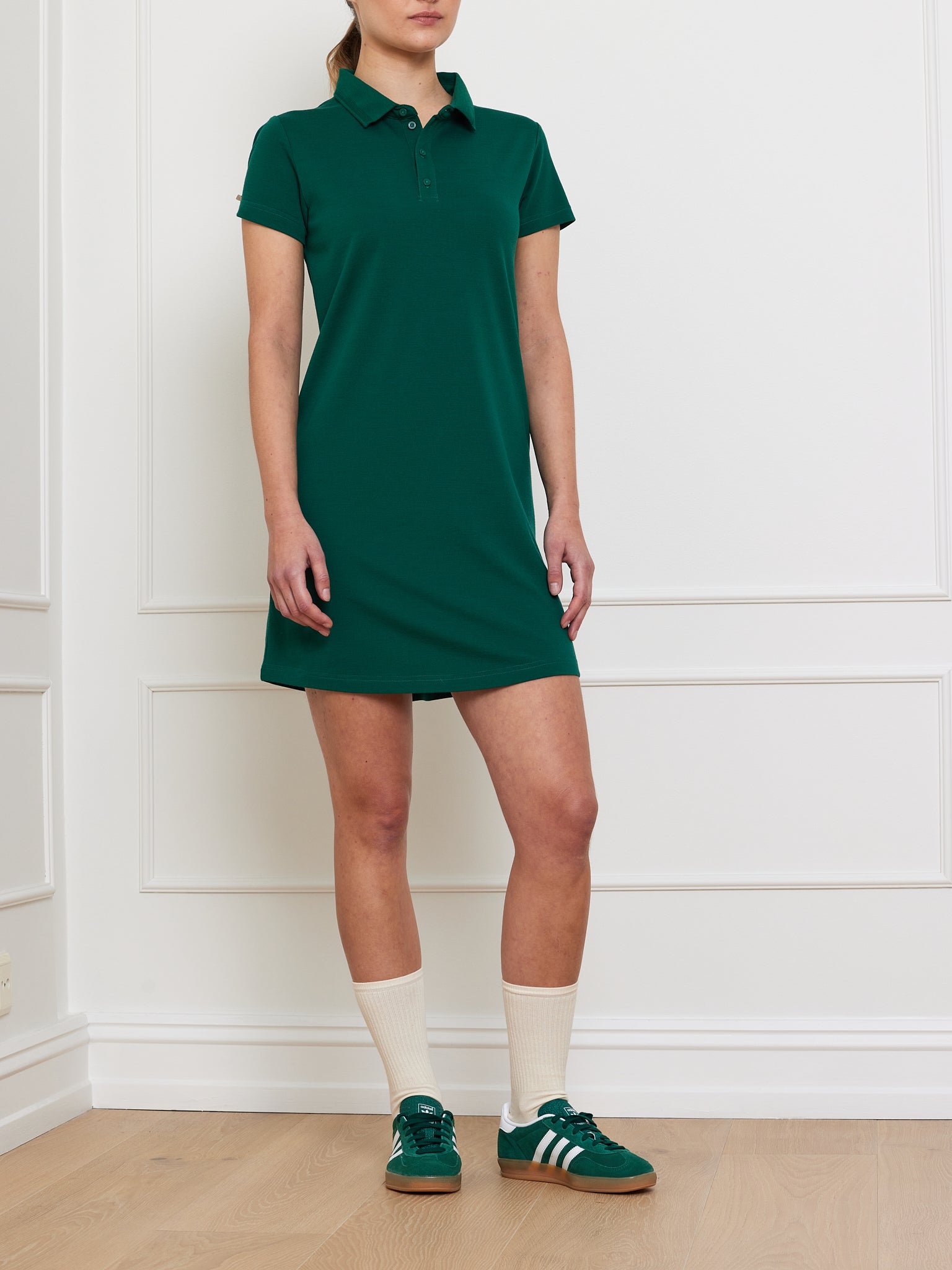 Indoors, a person wears a moisture-wicking Medalist Piqué Dress Women by We Norwegians—green with short sleeves—paired with cream knee-high socks and green sneakers. The stylish ensemble complements the light wood floor against the plain white wall perfectly.