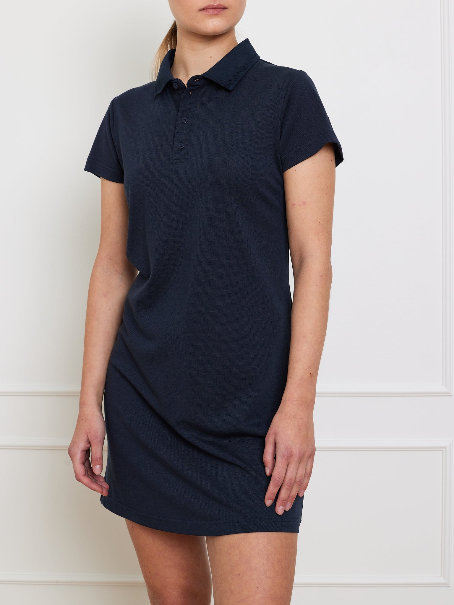 A person in a navy Medalist Piqué Dress Women by We Norwegians, made from moisture-wicking fabric with a button-up collar and relaxed fit, stands against a white wall. The image is cropped to display the upper body and upper legs.