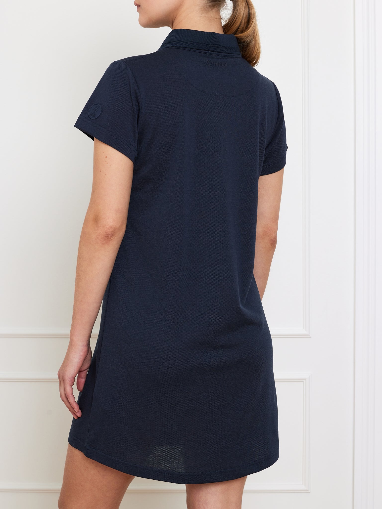 A person stands against a white wall, wearing a We Norwegians navy blue Medalist Piqué Dress Women with short sleeves. Their left arm rests by their side, and they have light brown hair tied back.