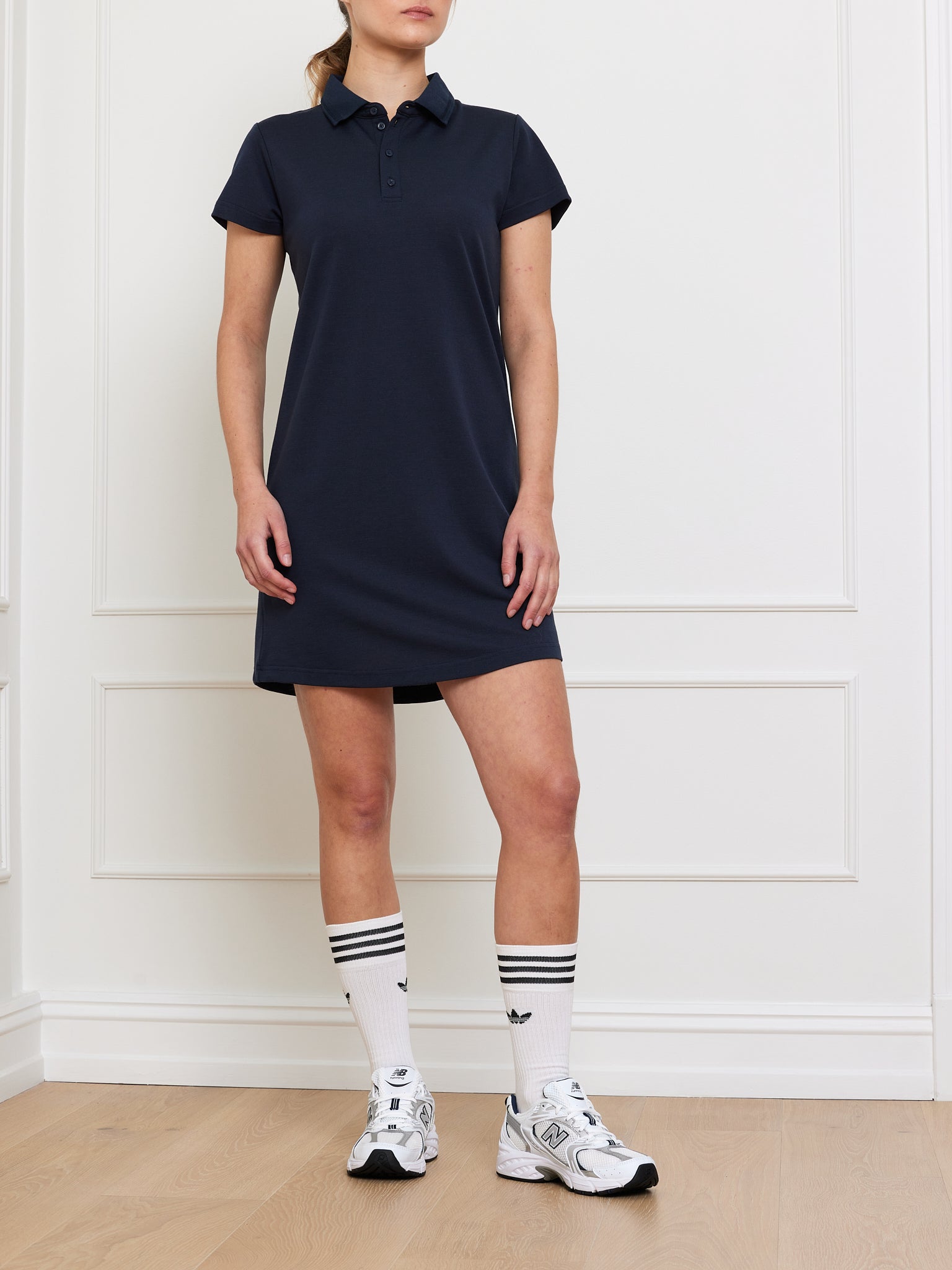 A person stands poised against a white-paneled wall on a light wood floor, donning a Medalist Piqué Dress Women from We Norwegians, paired with knee-high white socks featuring black stripes and crisp white sneakers.