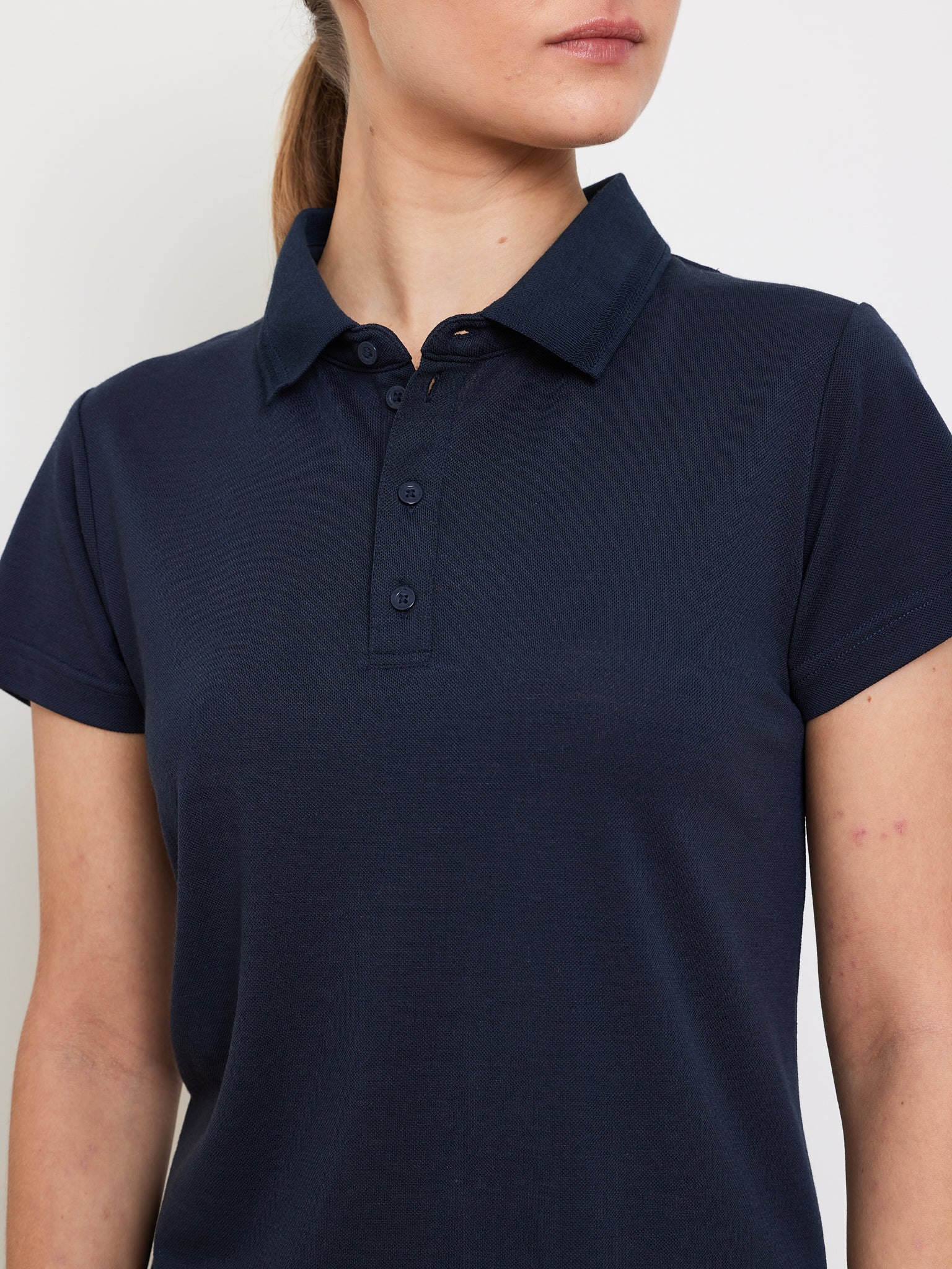 A person with light skin and long hair tied back wears the "Medalist Piqué Dress Women" by We Norwegians. The dark navy polo features a collar and button details, crafted from moisture-wicking fabric for all-day comfort, set against a plain background focusing on the upper body.