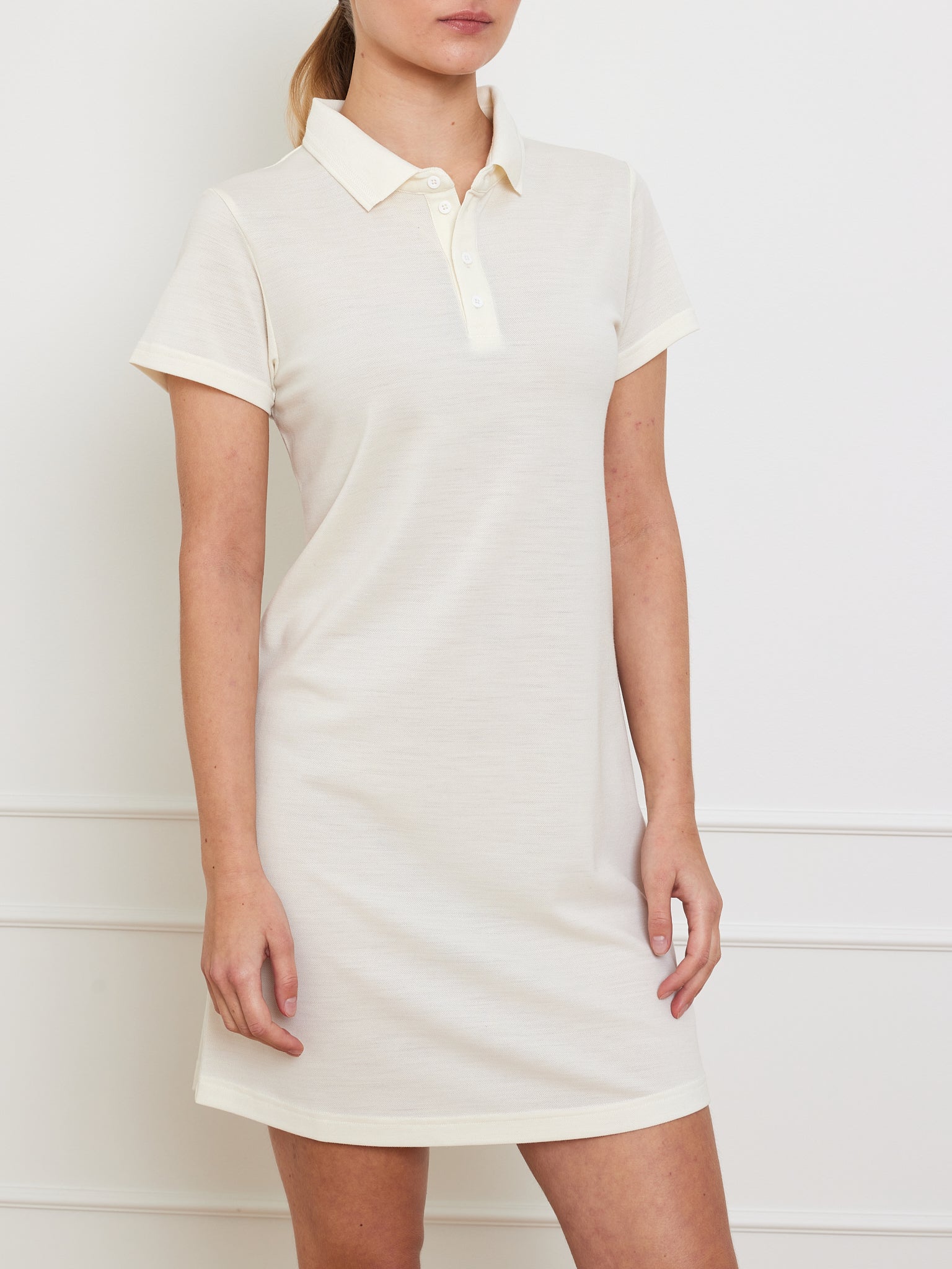 A woman in a short-sleeved, cream-colored Medalist Piqué Dress Women by We Norwegians stands against a blank white wall. The dress, featuring a classic collar and buttons, blends elegance with advanced moisture-wicking technology. Her face is not visible in the image.