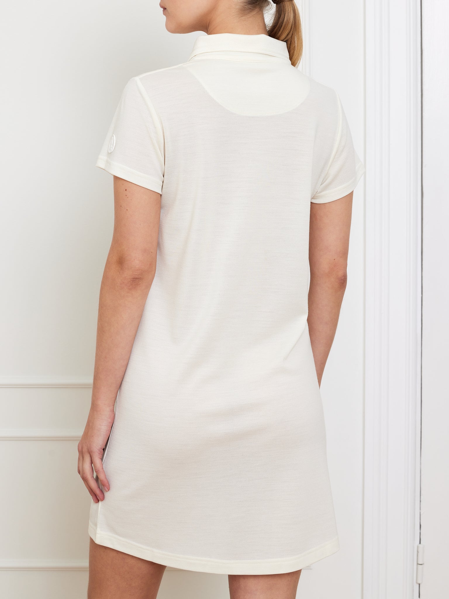 A person stands against a white wall, wearing the We Norwegians' Medalist Piqué Dress Women. The focus is on the back, highlighting its simplicity and elegance. Made from moisture-wicking fabric, the white short-sleeve collared dress blends style with functionality; their long hair is neatly tied back.