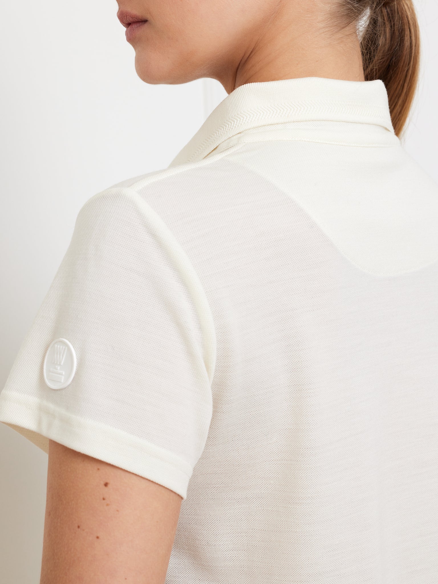 A person wears the Medalist Piqué Dress Women by We Norwegians, featuring a beige, short-sleeve polo design and a moisture-wicking logo on the sleeve. Their neat appearance is highlighted by a plain white background and a ponytail hairstyle.