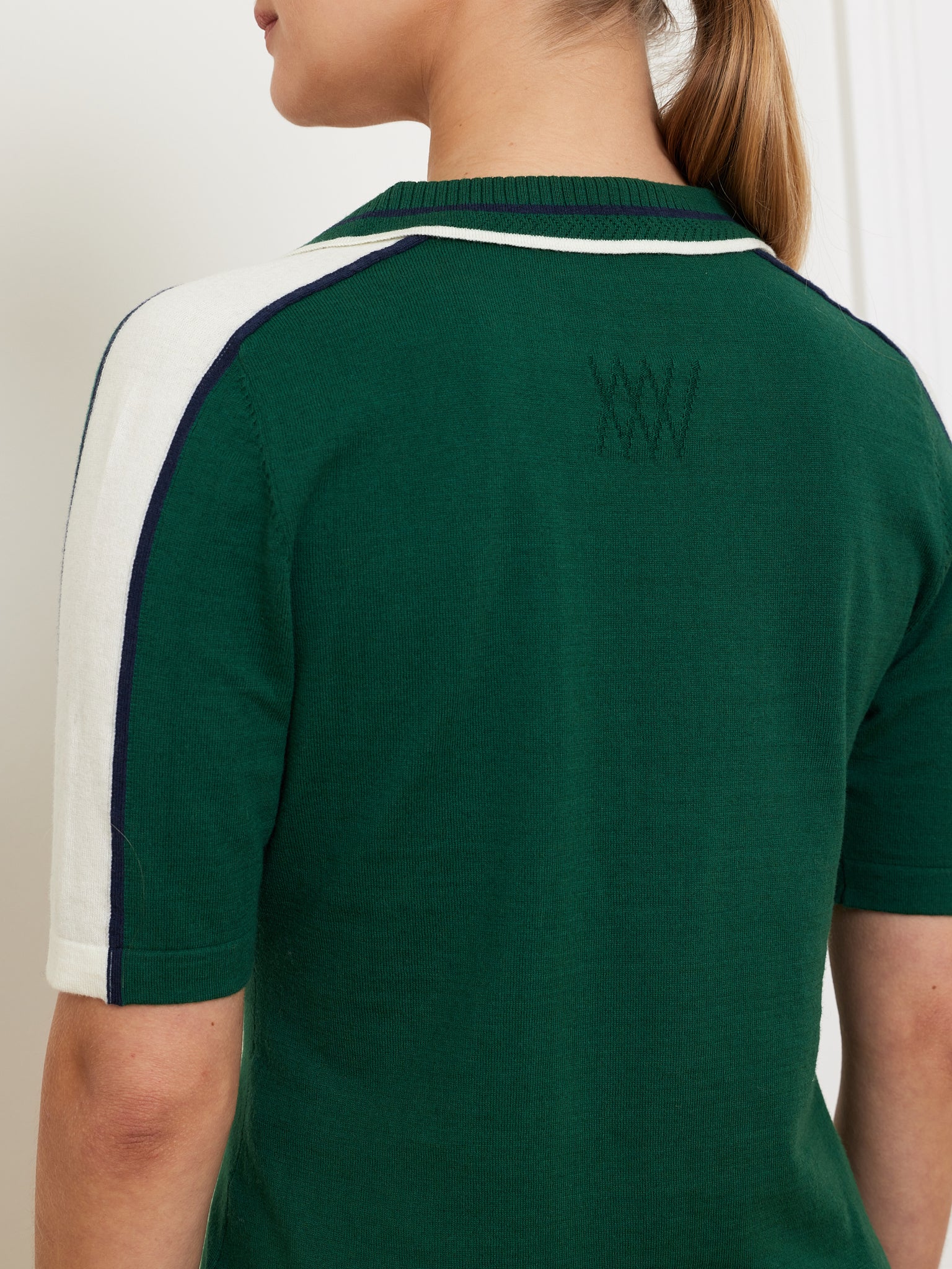 A person is seen from the back wearing a We Norwegians Team Polo Women in green, with white shoulder panels and dark trim featuring contrast piping. The ribbed collar complements the subtle embroidered design on the back, while their hair is pulled back.