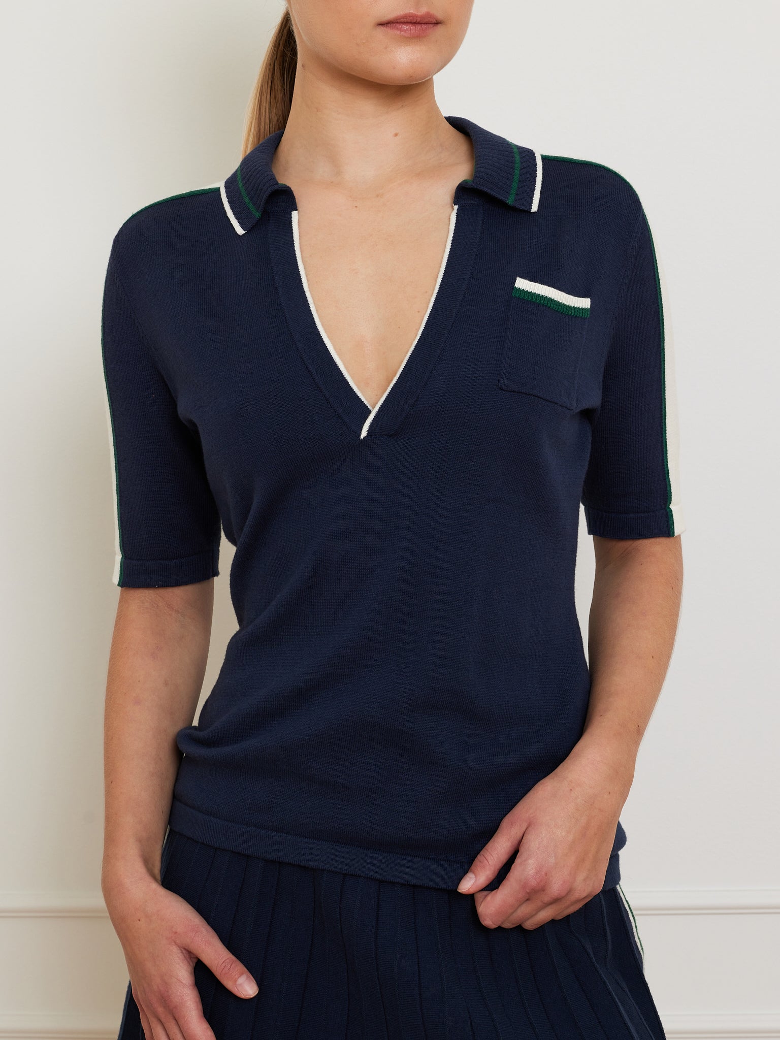A person wearing the We Norwegians Team Polo Women, a navy short-sleeve top with a deep V-neck, left pocket, and contrast piping on the collar and sleeves. Crafted from a soft merino-cotton blend, the wearer stands against a plain wall with hair tied back.