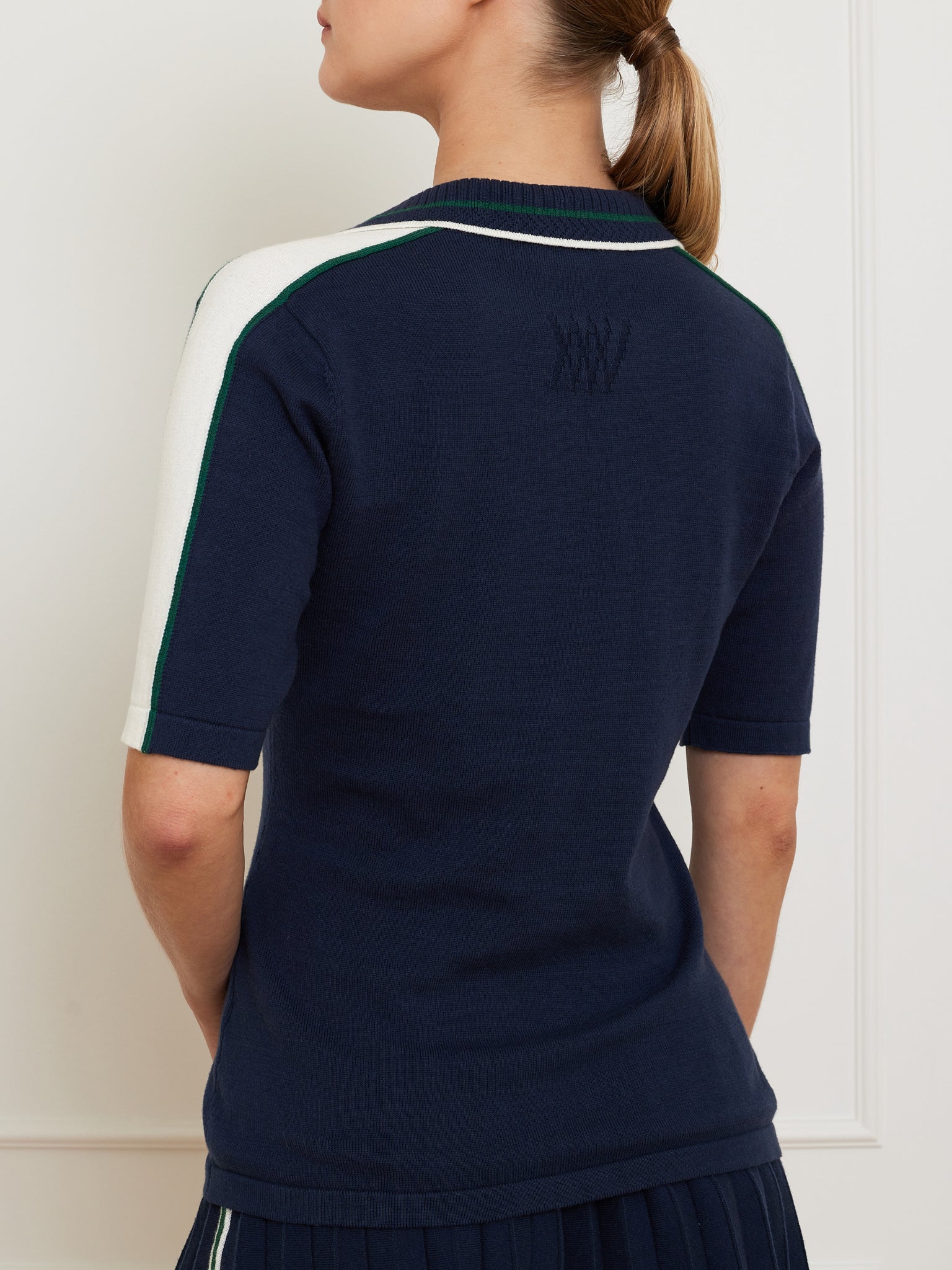 A person with a ponytail wears a navy blue Team Polo Women by We Norwegians, featuring a ribbed collar and white and green sleeve stripes. The merino-cotton blend shirt boasts subtle embroidery on the upper back. They are facing away from the camera.