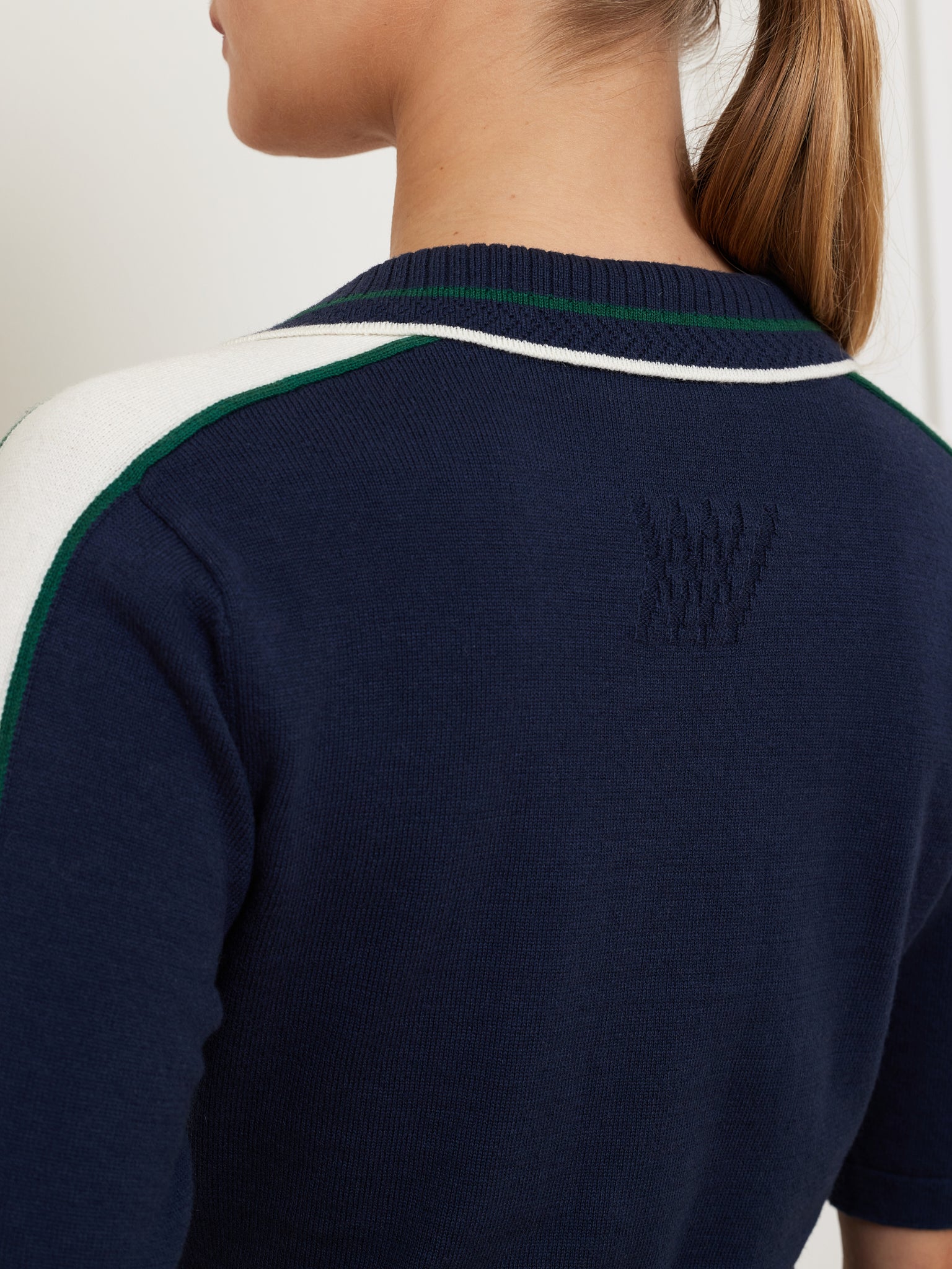 A woman seen from behind wears the We Norwegians Team Polo Women sweater in navy blue, featuring white and green striped sleeves. Her hair is styled in a low ponytail, highlighting the ribbed collar and subtle stitched logo on the back.