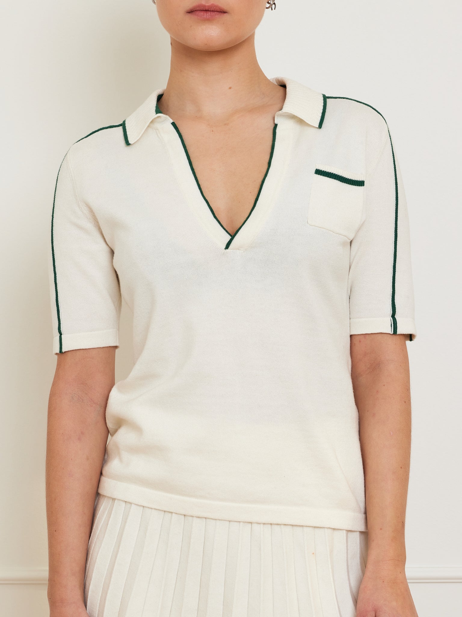 Dressed in We Norwegians’ Team Polo Women, a short-sleeve merino-cotton knit top with green piping on the V-neck and sleeves, paired with a white pleated skirt, the outfit stands out beautifully in the neutral-colored room.