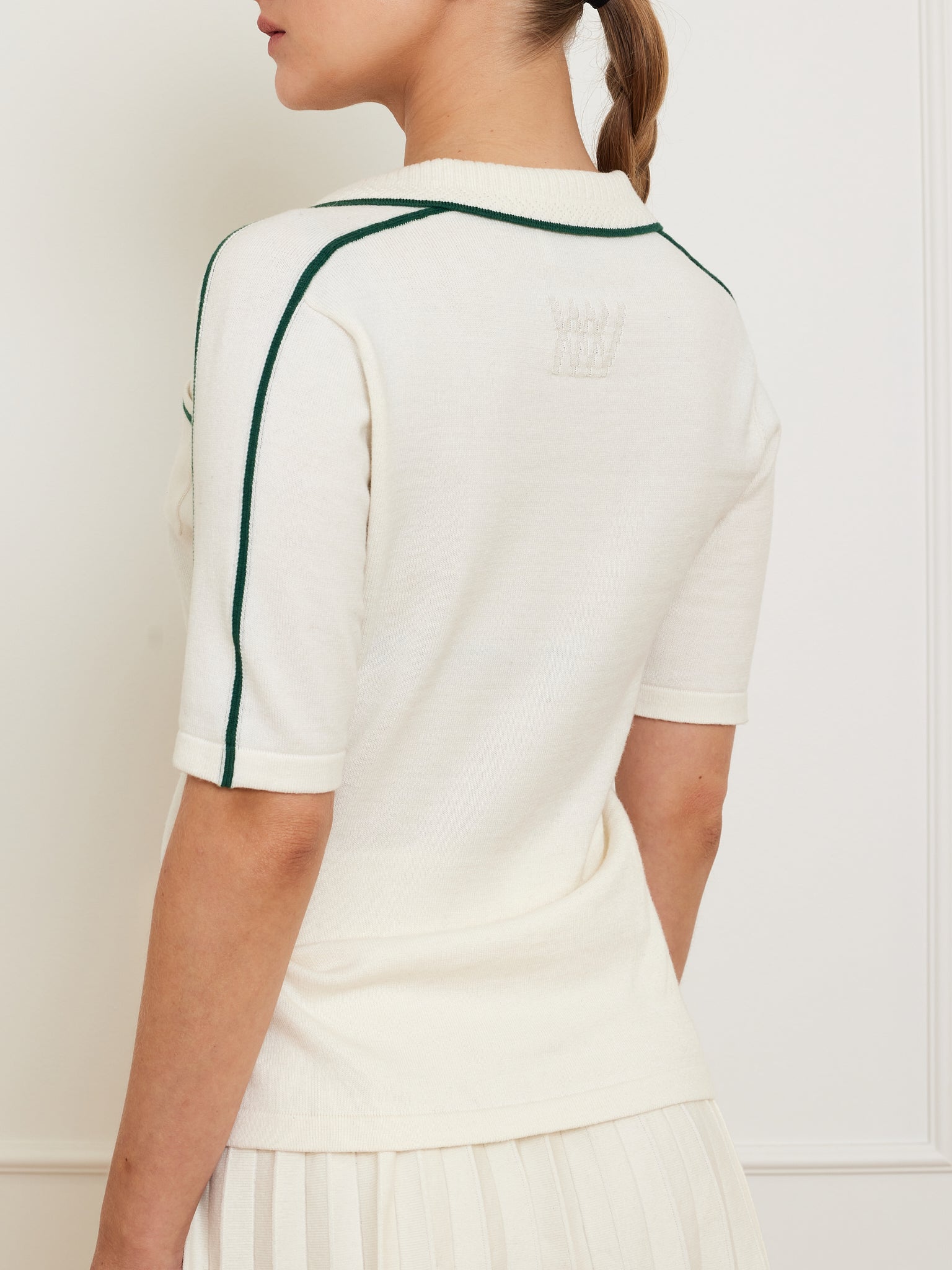 A person is wearing a We Norwegians Team Polo Women in white, featuring contrast piping on the shoulders, a ribbed collar, and a subtle emblem on the back, paired with a matching pleated skirt. Their hair is styled in a braid.