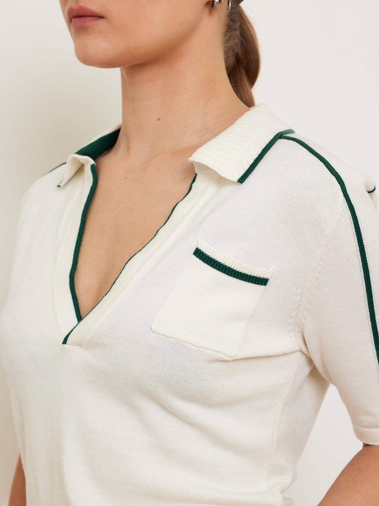A woman models the "Team Polo Women" by We Norwegians—a white short-sleeve shirt with a deep V-neck, green contrast piping, and chest pocket. It's crafted from a soft merino-cotton blend and set against a plain light-colored background.