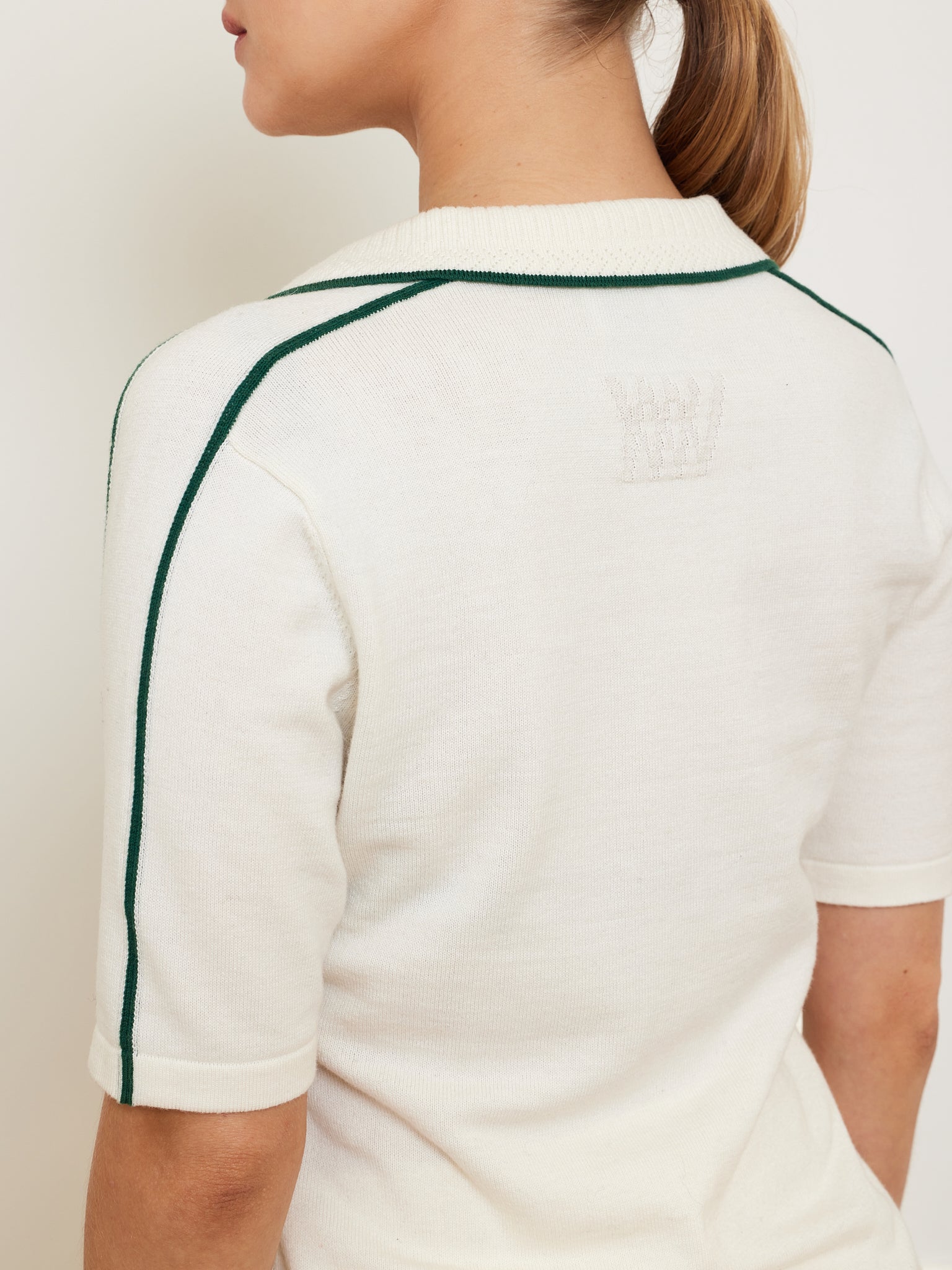 A person is wearing the We Norwegians Team Polo Women, a white merino-cotton blend short-sleeve top with green piping on the sleeves and an embroidered crown on the back. With their hair tied back, they face slightly away from the camera.