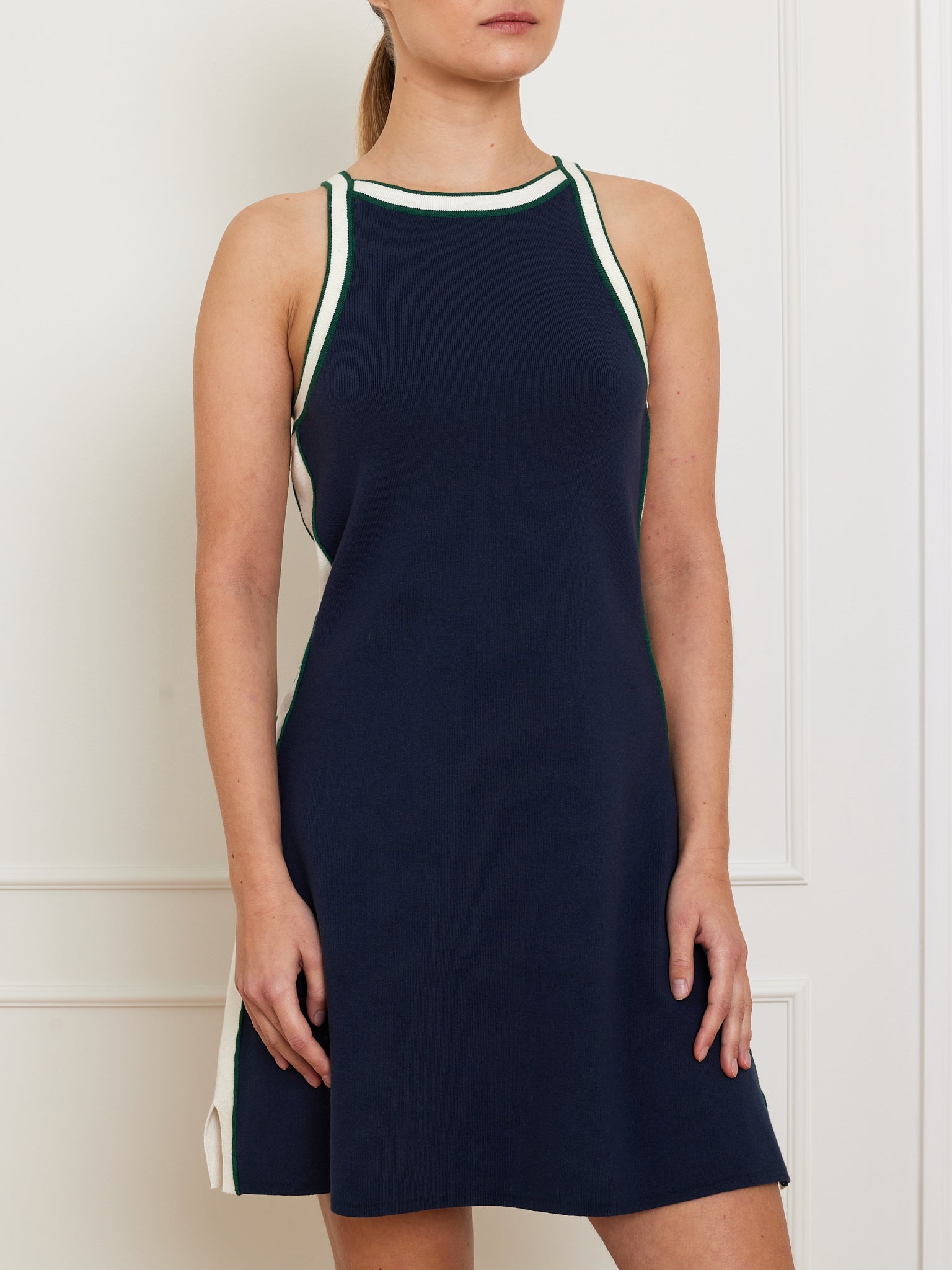 A person models the Team Dress Women by We Norwegians against a white wall. This sporty, navy blue dress features body-contouring panels, a racerback design, and sleeveless style with green and white trim along the neckline and sides, adding a simple yet chic flair.
