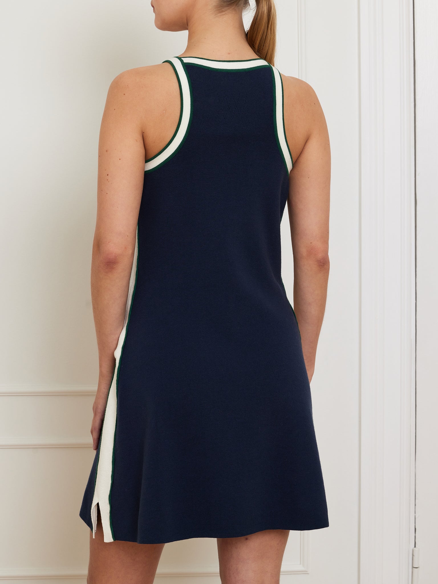 Someone wears the We Norwegians Team Dress Women, a sleeveless navy blue merino-cotton blend dress with a racerback design, white and green trim, and a side slit. They stand with their back to the camera against a white wall.