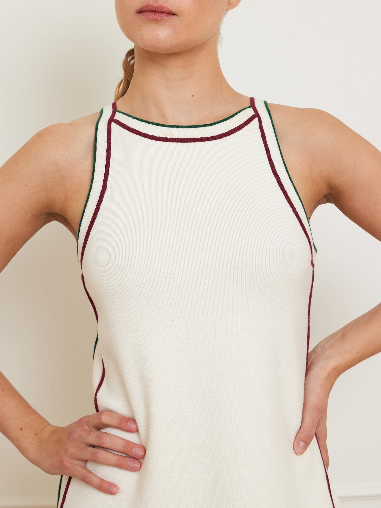 A woman models the "Team Dress Women" by We Norwegians, showcasing its sleeveless white design with green and red edging, and a racerback style, while standing with arms akimbo.