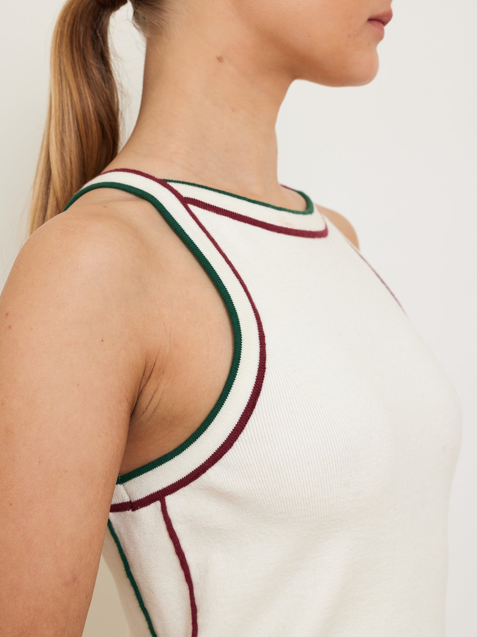 A person with long hair tied back is wearing the We Norwegians Team Dress Women, a white sleeveless top with red and green piping and a sleek racerback design. The focus is on the upper torso and neck against a plain background, highlighting the dress's stylish cut.
