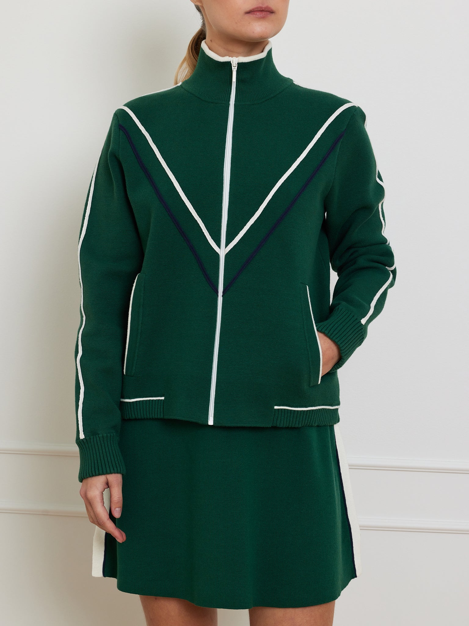 A person wears the Team Jacket Women by We Norwegians, a dark green retro-style jacket with white accents and a V-shaped design, long sleeves, a zip front, and paired with a matching skirt. They casually place their right hand in the pocket while standing against a neutral background.