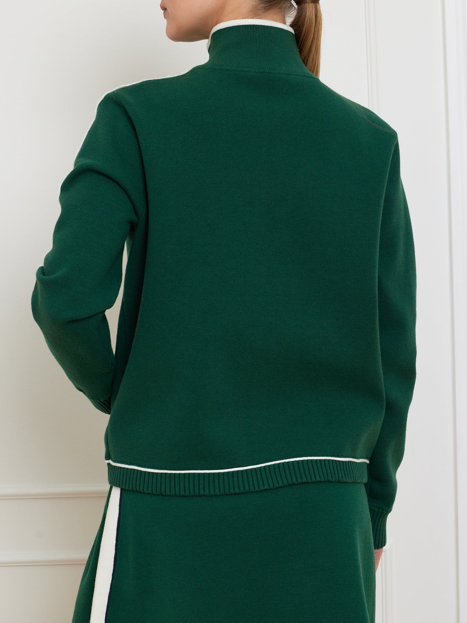 A person stands with their back to the camera, wearing We Norwegians' Team Jacket Women in forest green with white trim and a matching green skirt featuring a white side stripe. The ensemble creates a retro track jacket vibe, and they have their hair in a ponytail.