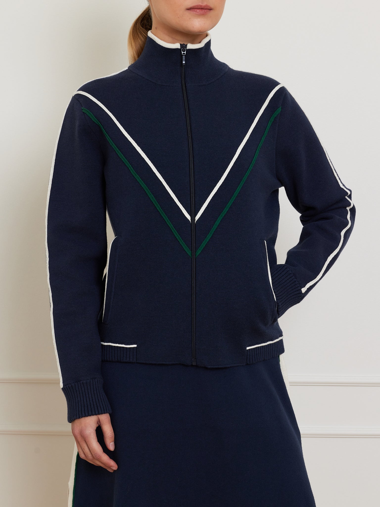 A person models We Norwegians' Team Jacket Women, a retro navy blue jacket with green and white V-shaped stripes, made of a cotton-merino blend. It has a high collar and ribbed cuffs and is paired perfectly with a matching navy skirt against a plain, light-colored wall.