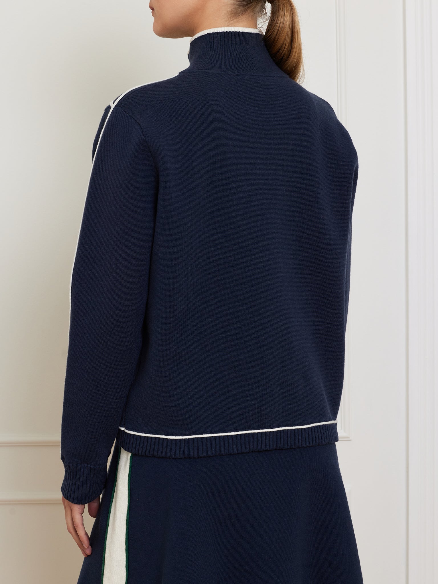 A person wearing We Norwegians' Team Jacket Women, a navy turtleneck sweater with white trim and a matching skirt for a relaxed fit, faces away from the camera in a minimalist interior setting.