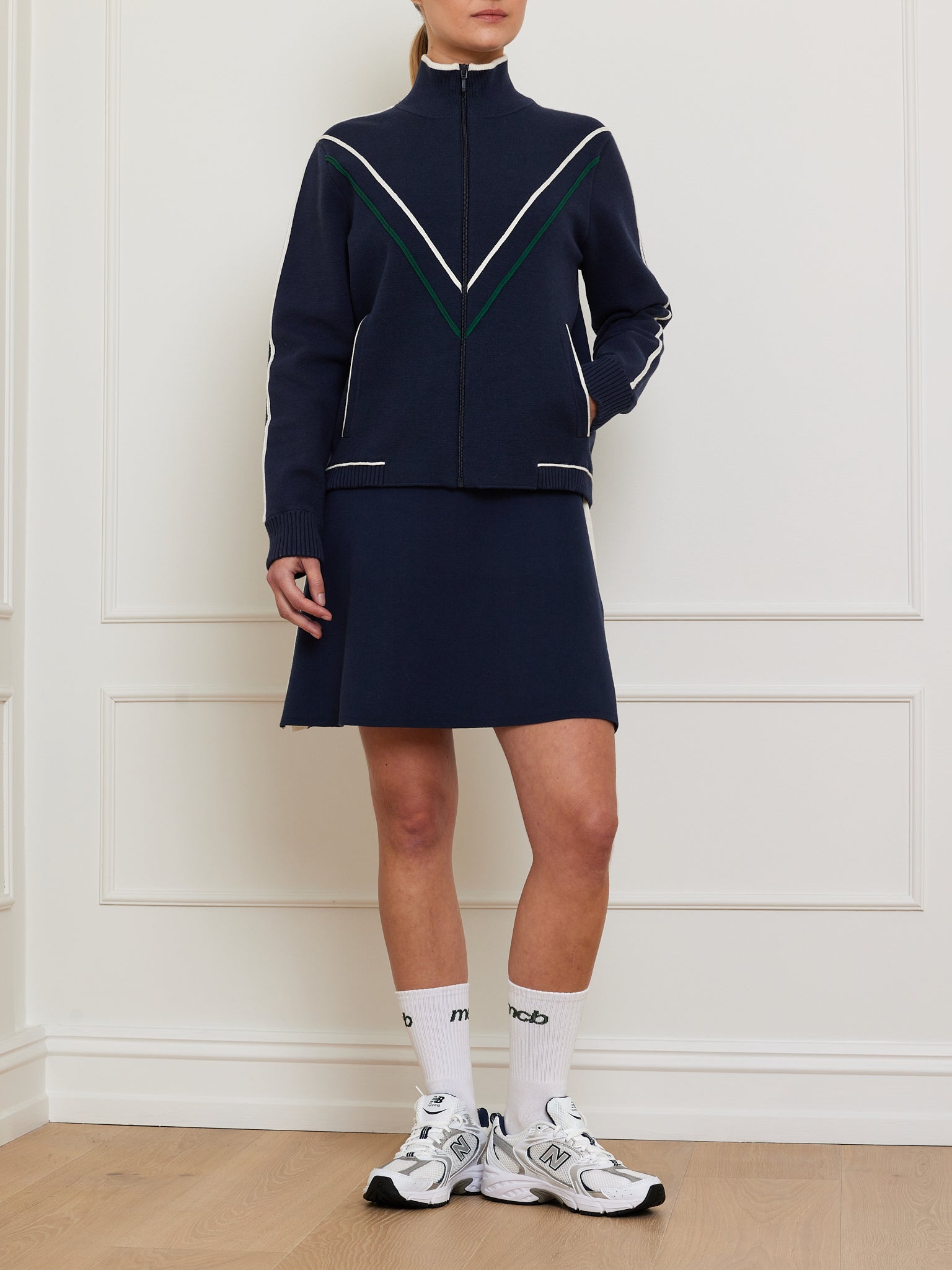 A person poses by a white paneled wall wearing We Norwegians Team Jacket Women, a navy retro track jacket with white and green chevron details, paired with a matching relaxed-fit skirt. The ensemble is completed with text-adorned white calf-length socks and crisp white sneakers.