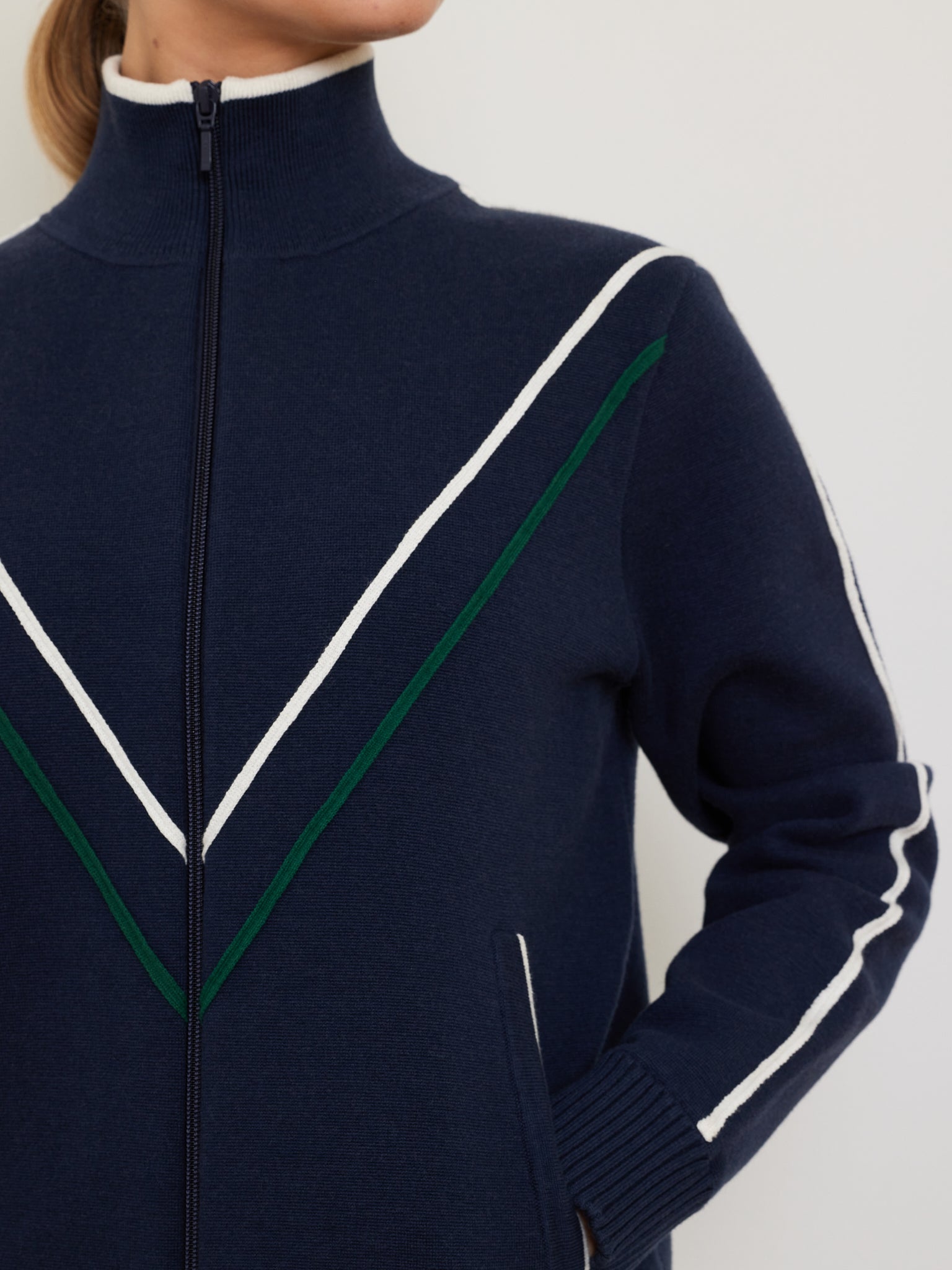Wearing the Team Jacket Women by We Norwegians, a person sports a retro navy track jacket with a high collar, featuring white and green V-shaped stripes and white arm stripes. The relaxed-fit design leaves their face hidden.