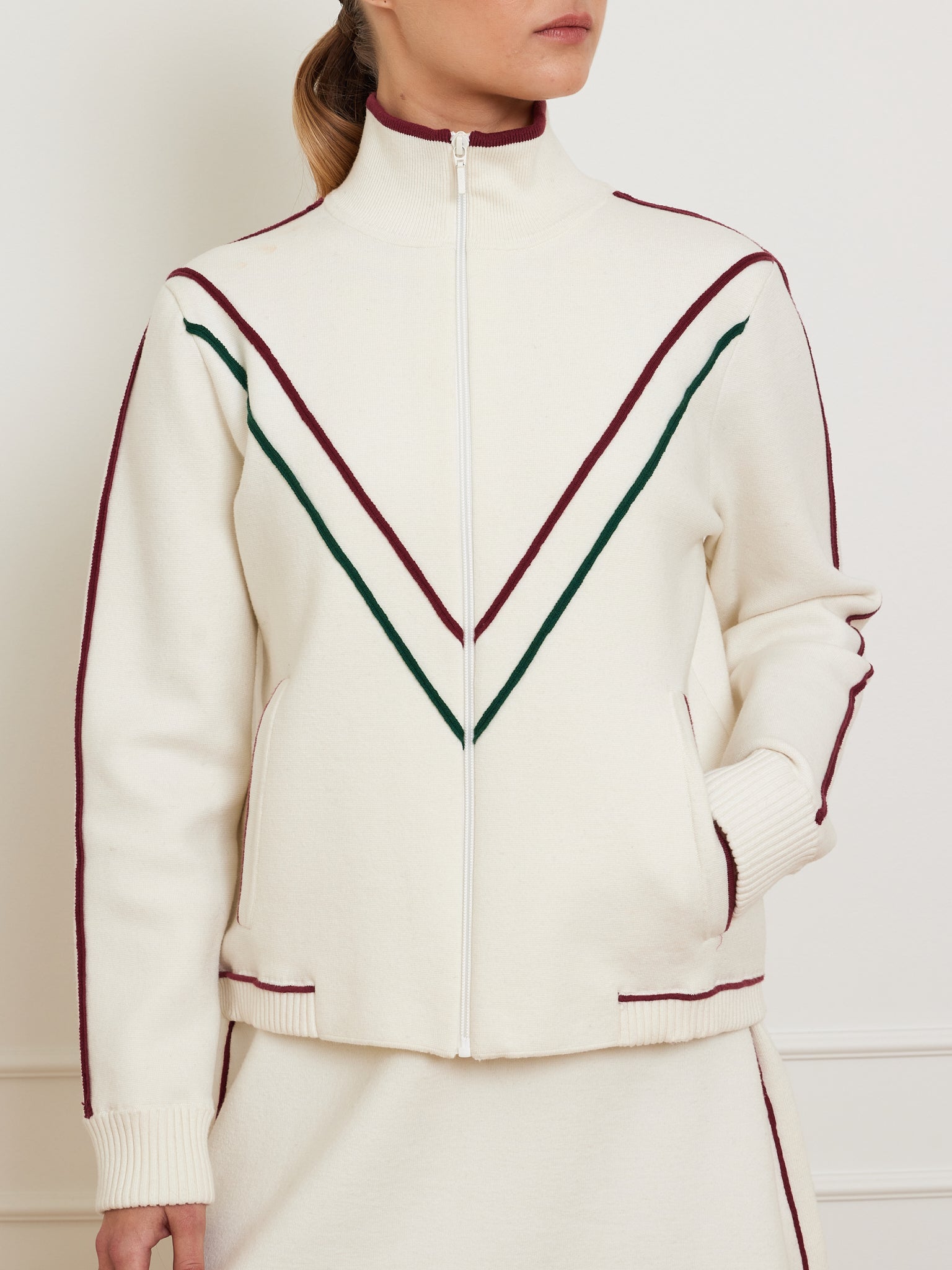 A person dons the Team Jacket Women by We Norwegians, a retro cream track jacket with burgundy and green V-shaped chest stripes. Made from a cotton-merino blend, it features burgundy piping on sleeves and pockets. Zipped up against a plain backdrop, it offers a relaxed fit.