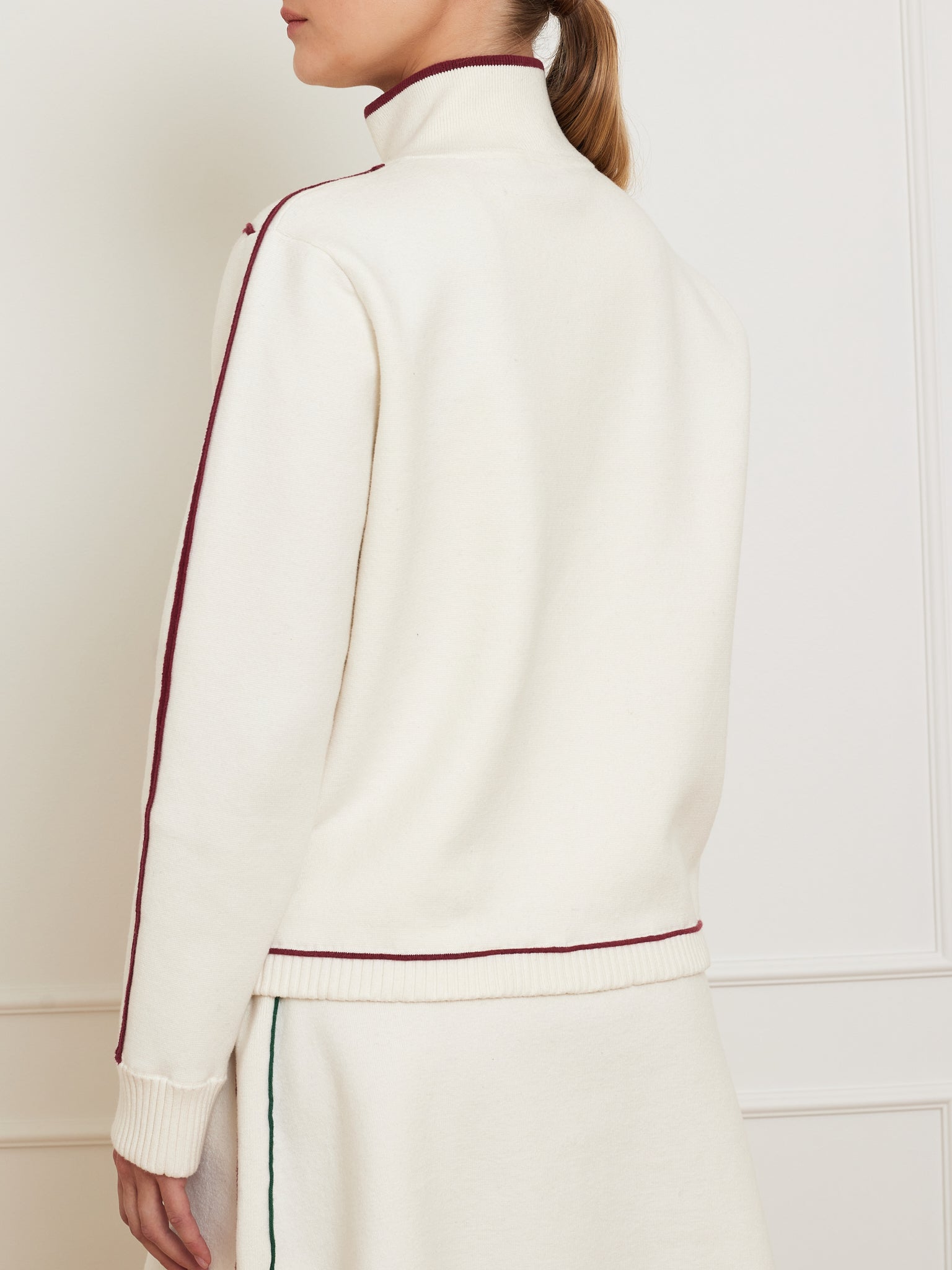 A person models We Norwegians' "Team Jacket Women," a cream outfit with a relaxed fit sweater and matching skirt. The cotton-merino blend sweater, featuring maroon piping along shoulders and sides, is turned slightly to display its stitching detail.