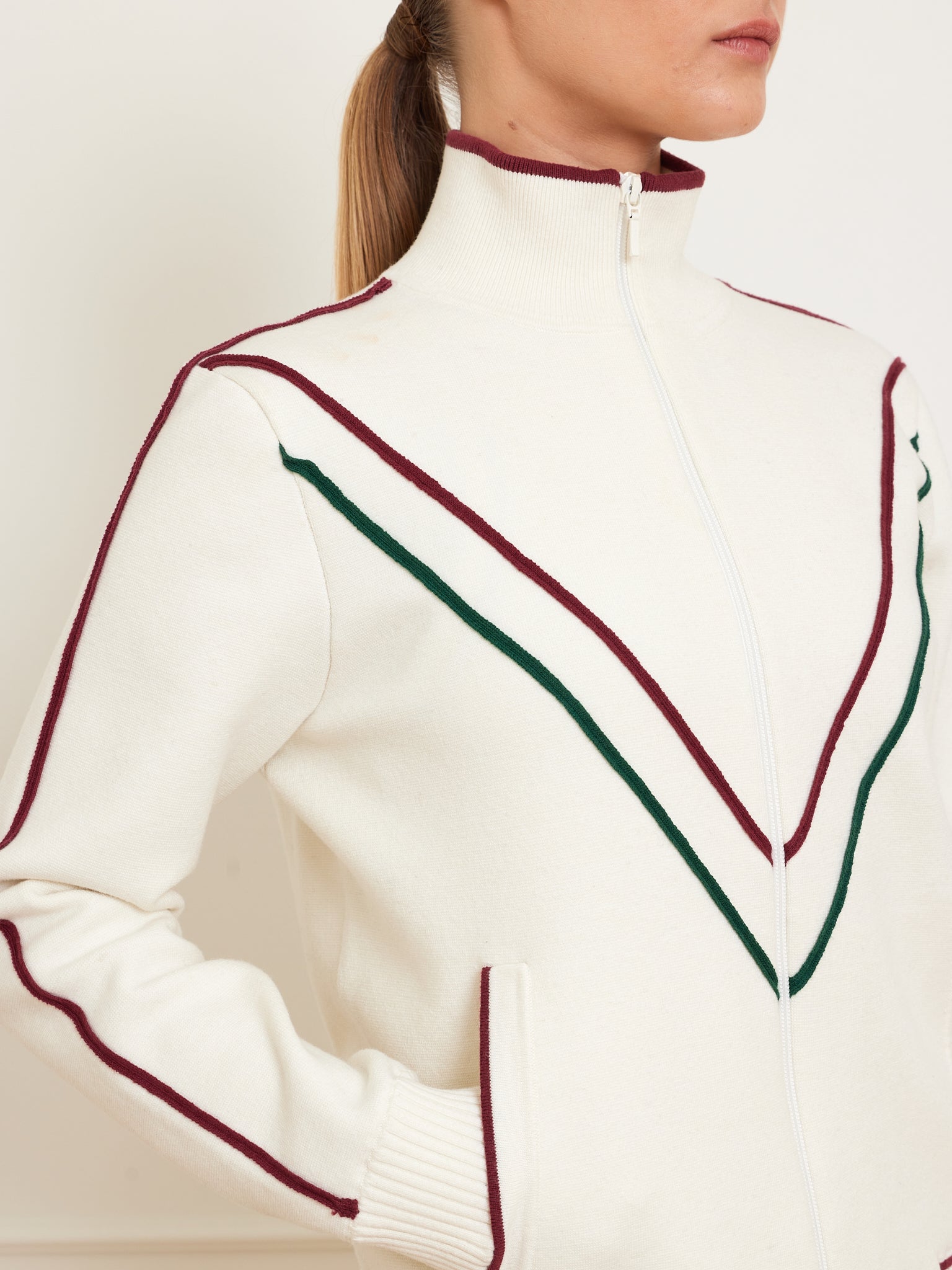 Wearing a We Norwegians Team Jacket Women, featuring red and green chevron stripes and a high collar, a person with tied-back hair shows only their lower face against a light background.