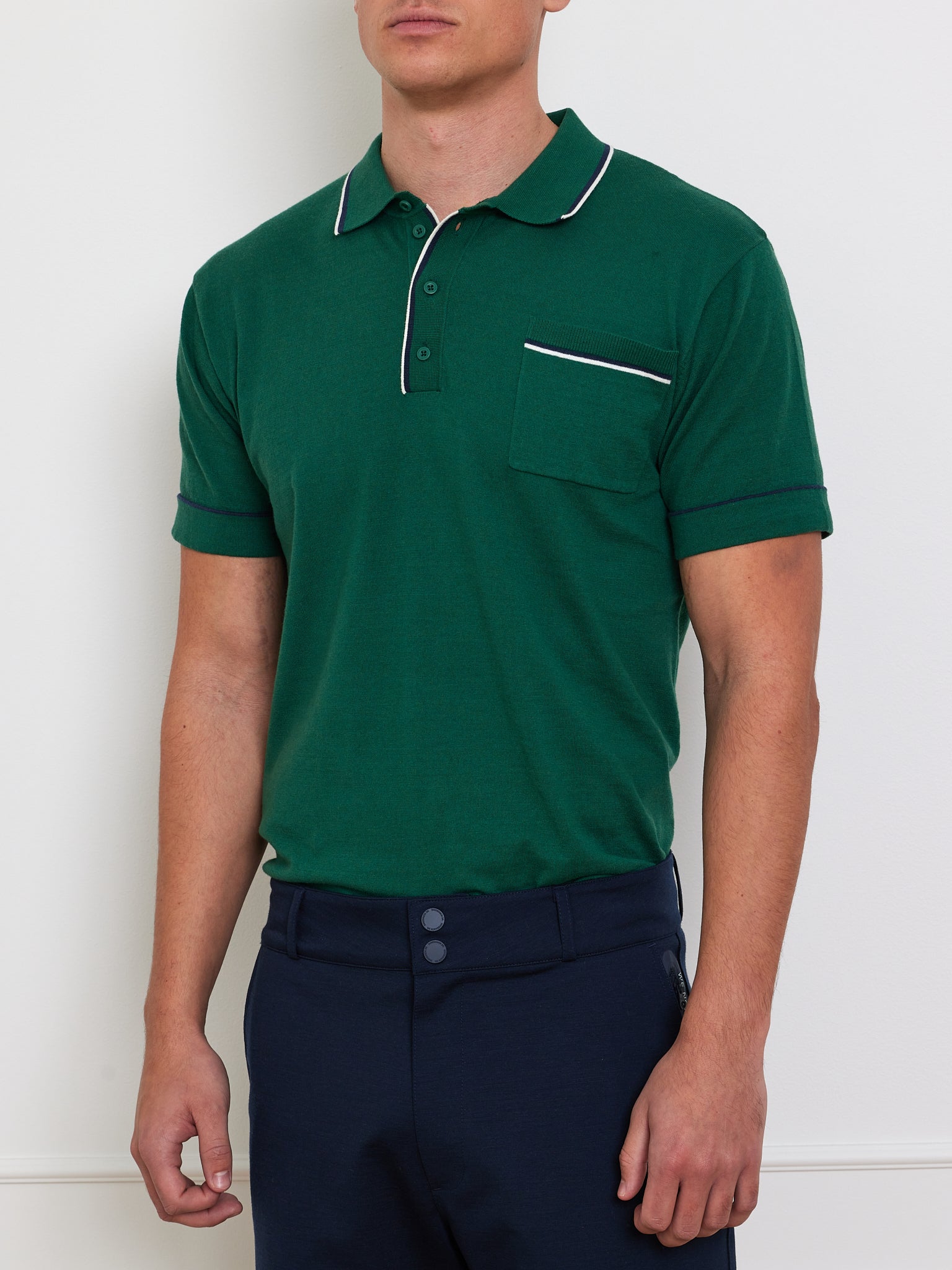 A person wears the "Team Polo Men" by We Norwegians, crafted with breathable fabric in green with white trim, featuring a chest pocket and buttoned collar. They pair it with dark navy pants against a plain white background.