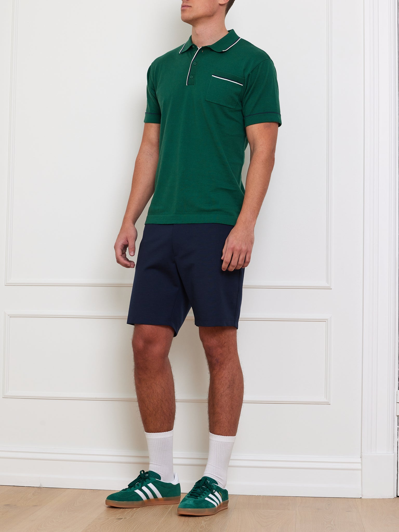 A person stands against a white wall wearing We Norwegians' Team Polo Men in green with white trim, navy shorts, and green sneakers with white stripes. White socks are visible on the wooden floor. The background is minimalistic.